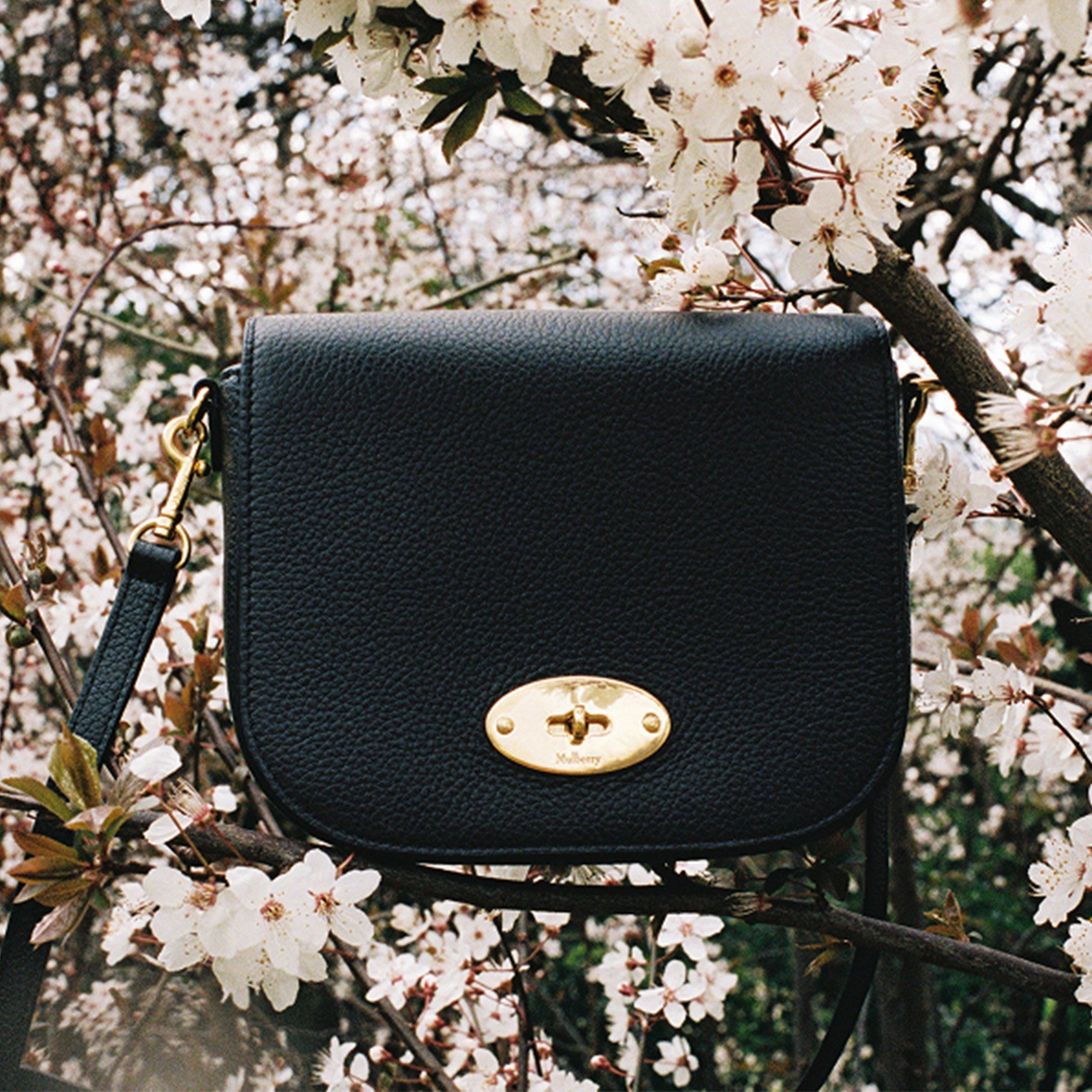 A black Mulberry crossbody bag with gold hardware hanging among blooming white flowers