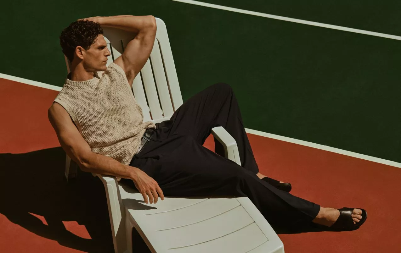 Male model lounging on sunbed at tennis court, wearing sleeveless knit top, black trousers and sandals