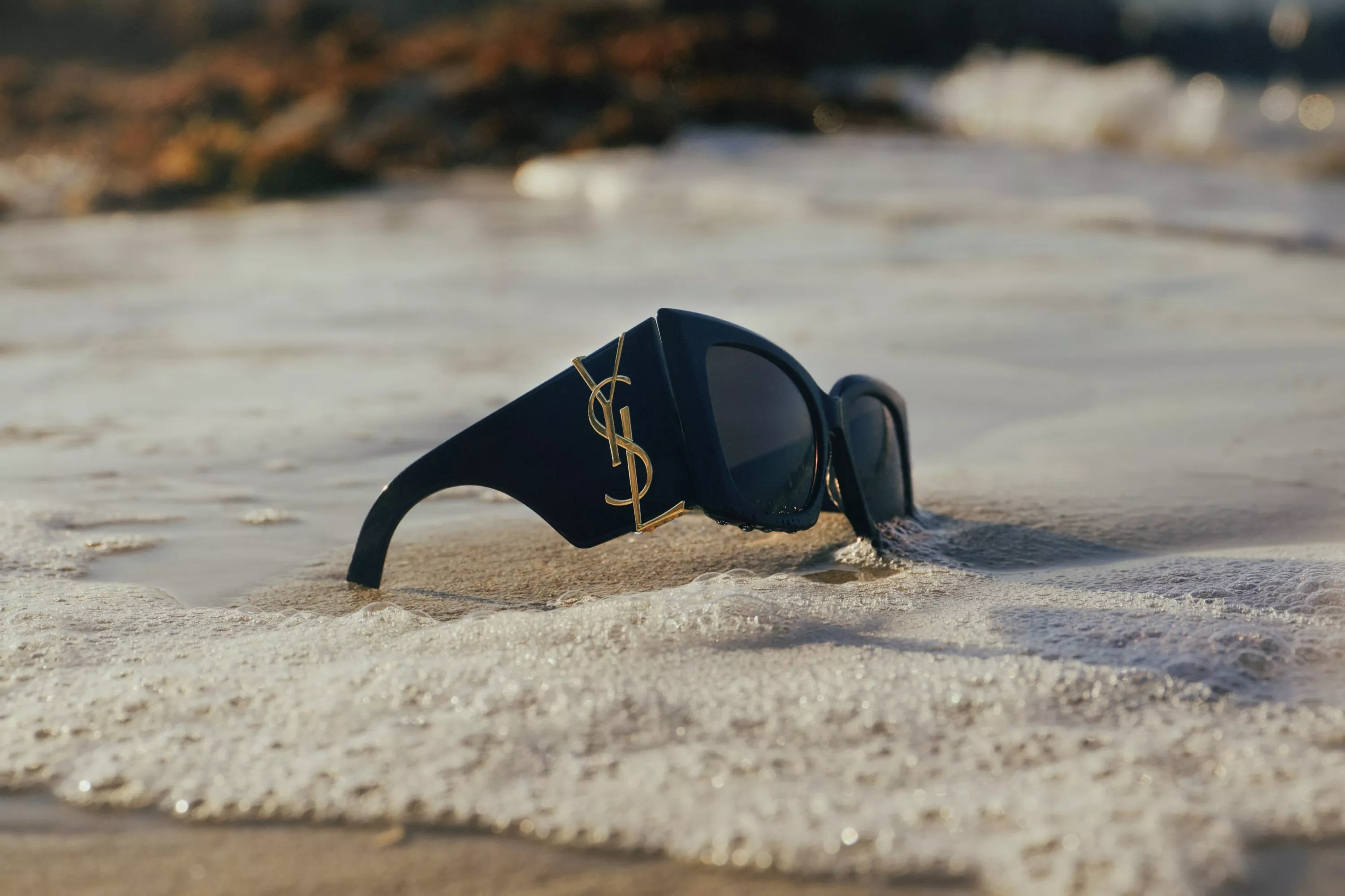 Black designer sunglasses washed by sea foam on sandy beach, luxury summer fashion accessory close-up