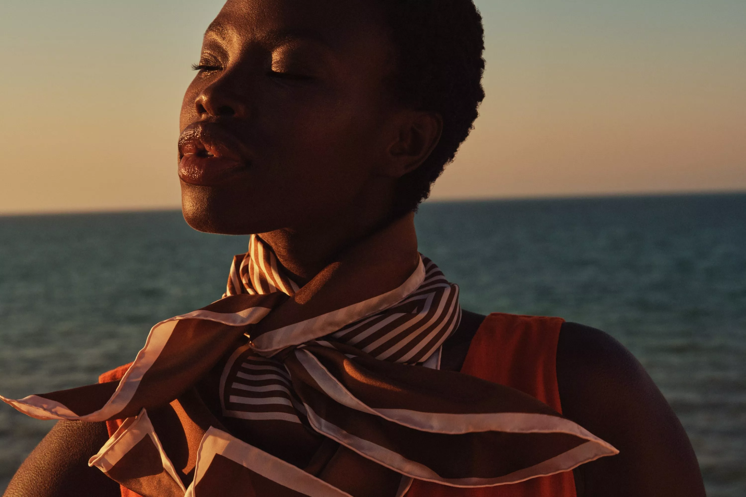 Close-up portrait of a woman by the sea at golden hour, wearing a striped scarf and orange top, eyes closed.