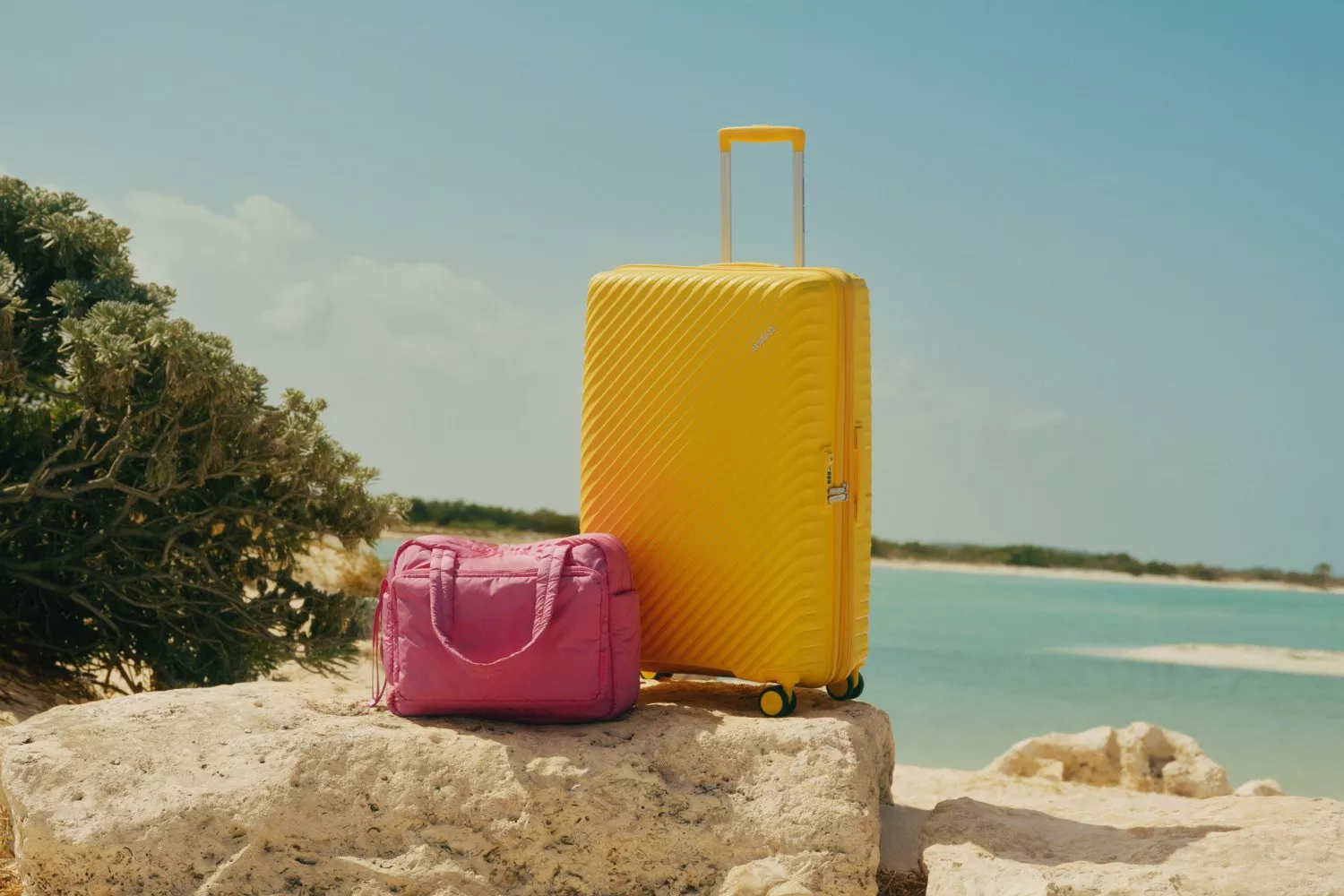 Yellow hard-shell suitcase and pink travel bag on rocks by a sunny beach lagoon, summer holiday luggage concept