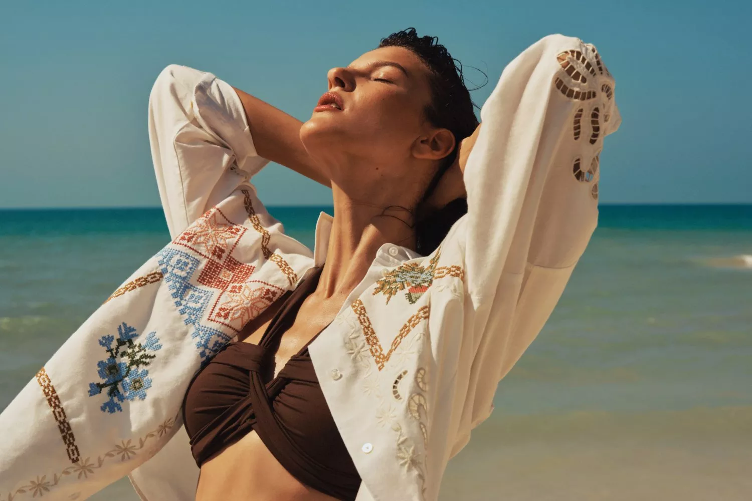 Woman in embroidered white beach shirt over brown bikini, posing by the sea in summer sunshine, travel fashion photo