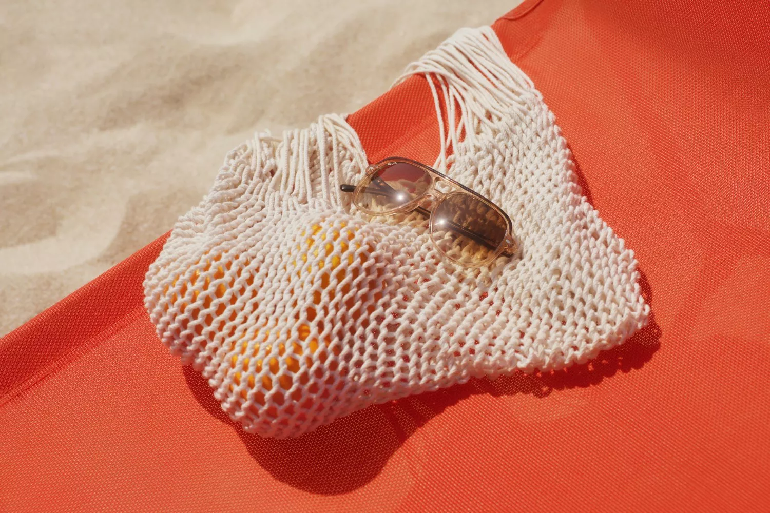 White crochet beach bag with sunglasses and citrus fruit on an orange towel in bright summer sunshine