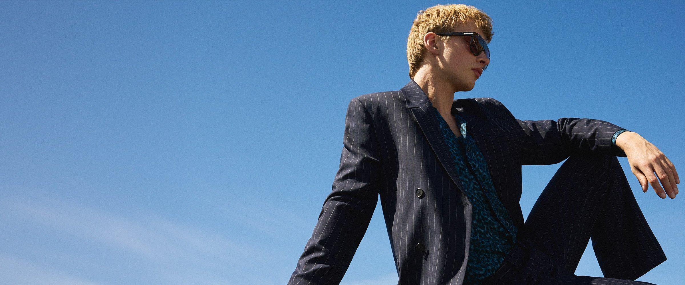 A man wearing a dark pinstripe suit and sunglasses poses confidently against a clear blue sky
