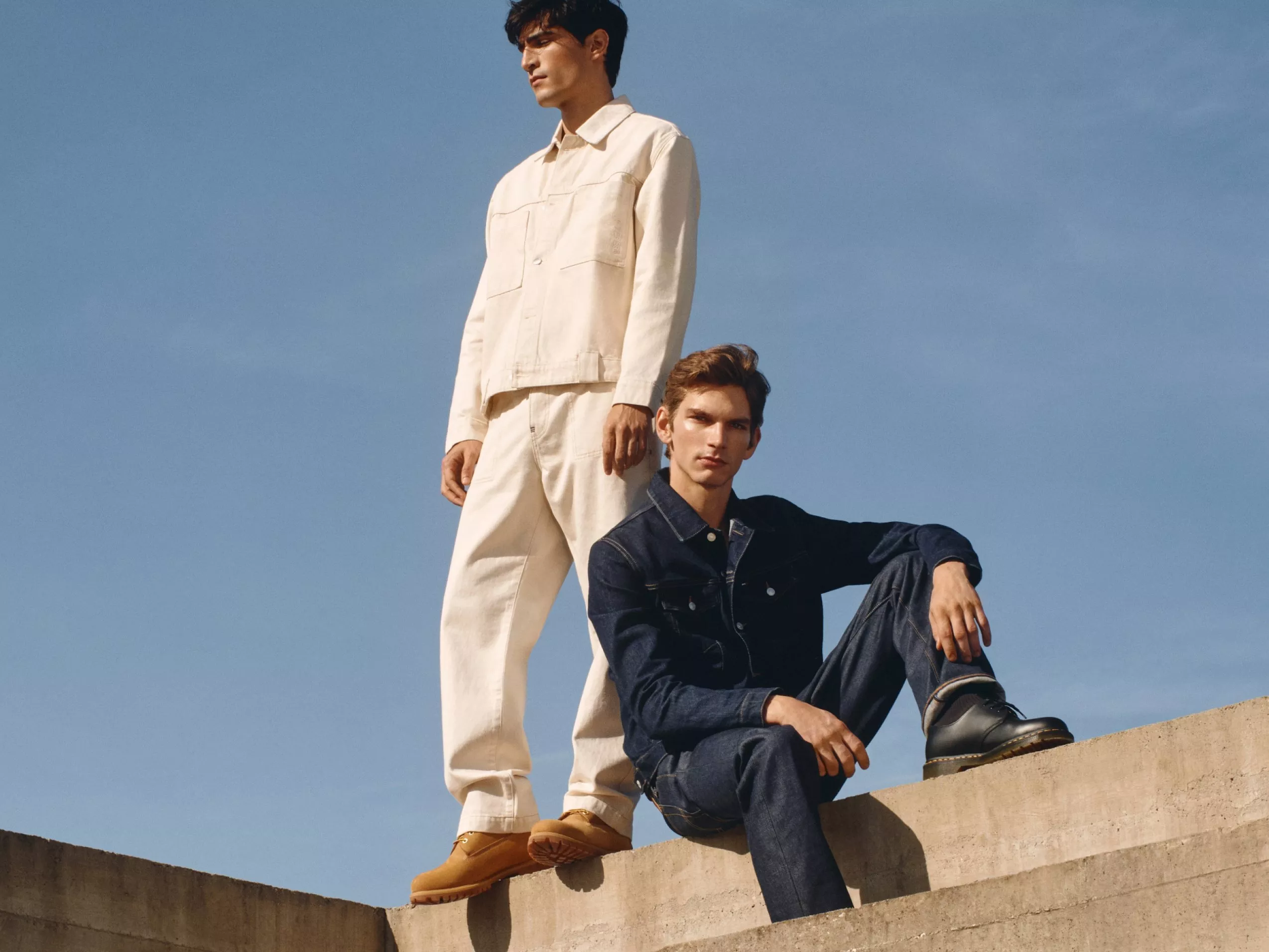 Two individuals wearing stylish jackets and trousers on a concrete structure under a clear blue sky.