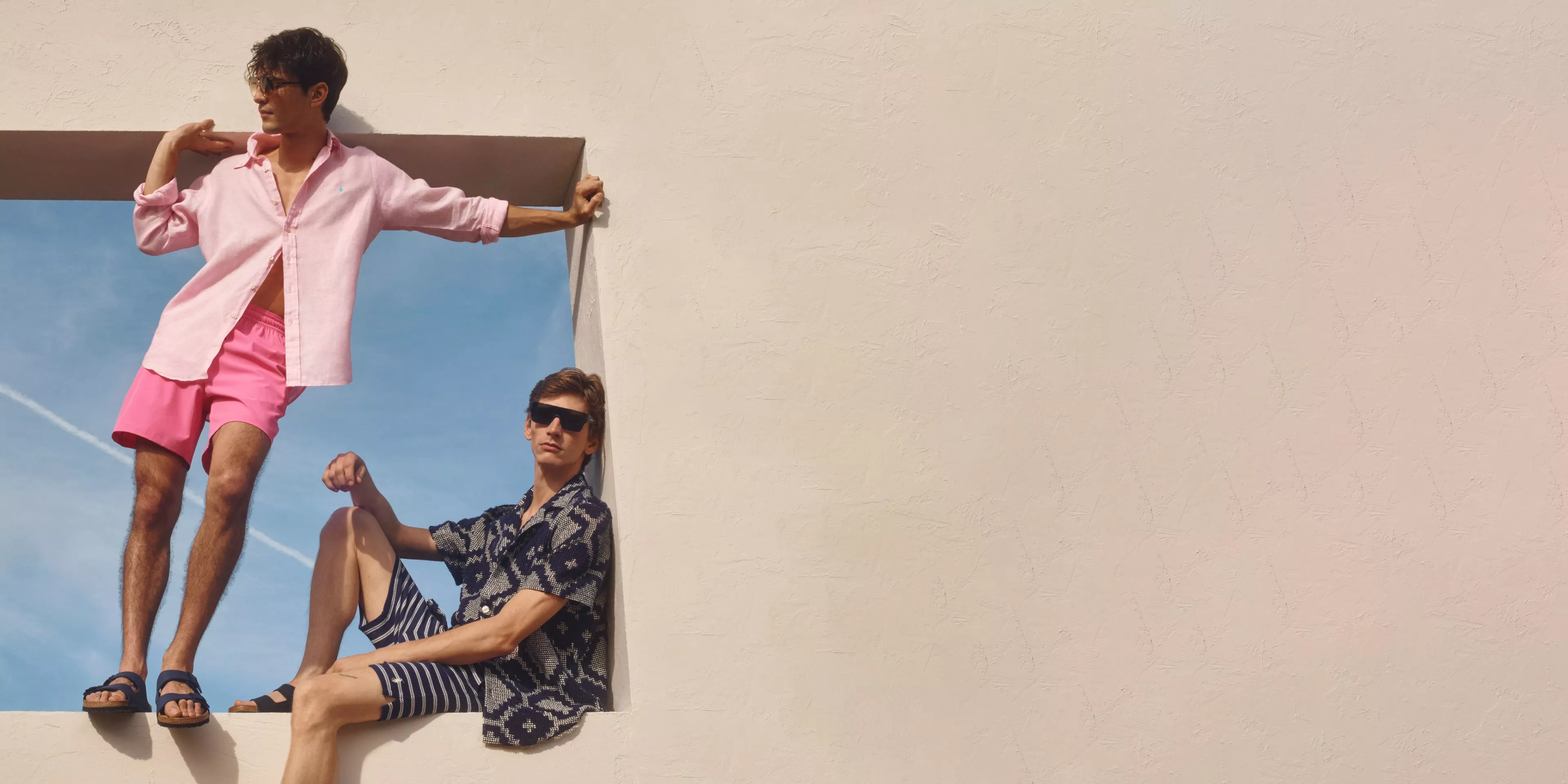 Two men in casual summer outfits pose in a window frame against a clear blue sky, exuding a relaxed vibe.