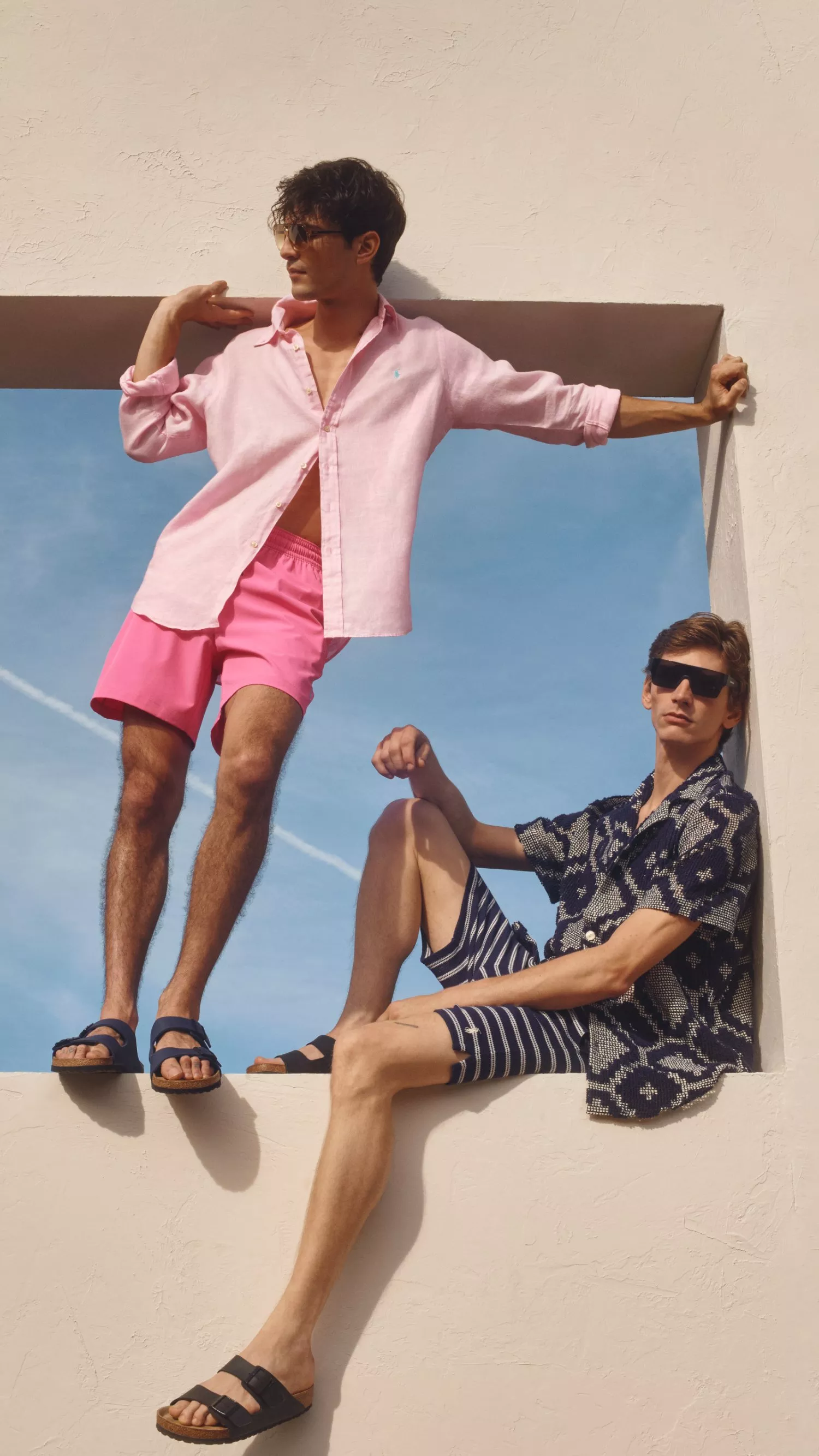 Two men in summer outfits, one in pink and the other in patterned navy, pose stylishly on a wall under a blue sky.