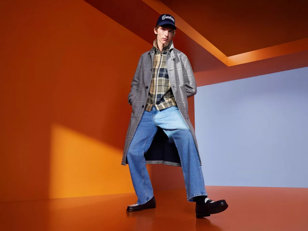 Men’s fashion model in long checked coat, plaid shirt and blue jeans, posing in modern orange studio set