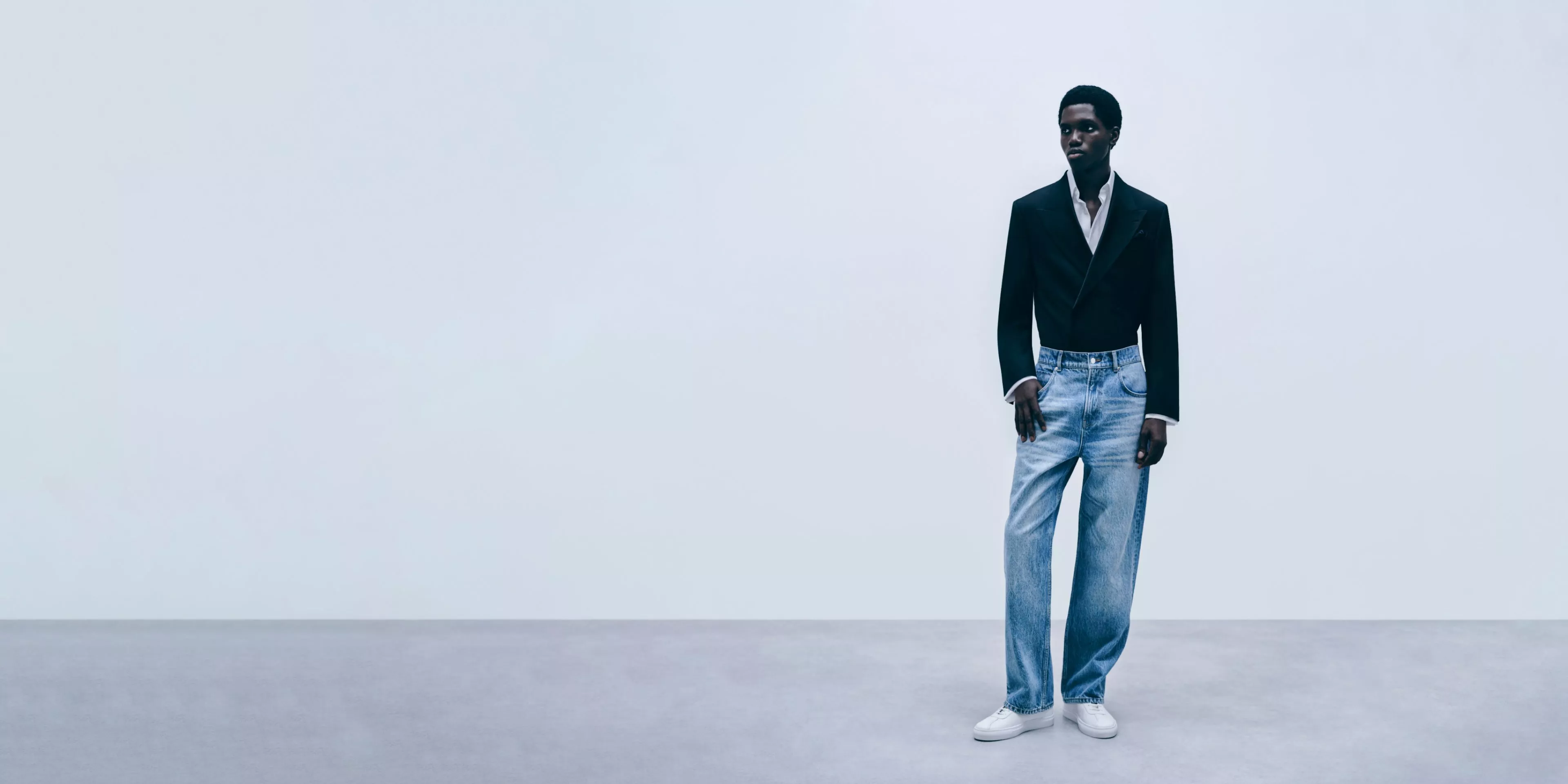 Men’s fashion model in black blazer, white shirt and light blue jeans, standing in a minimalist studio backdrop