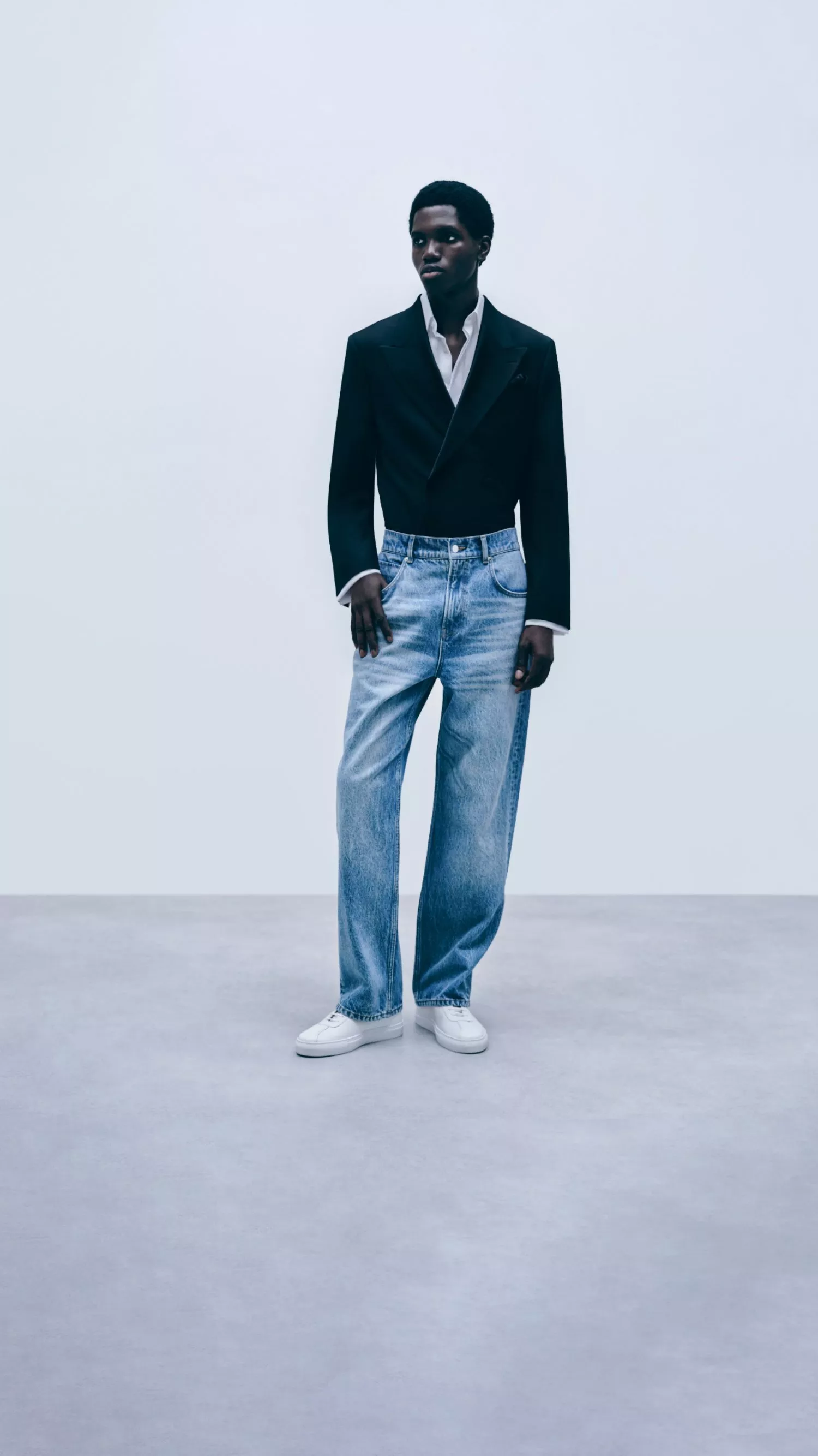 Full-length studio photo of male model in black blazer, white shirt and wide-leg jeans on pale backdrop