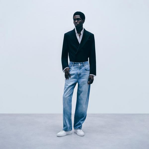 Male fashion model wearing black blazer, white shirt and light blue jeans in minimalist studio setting