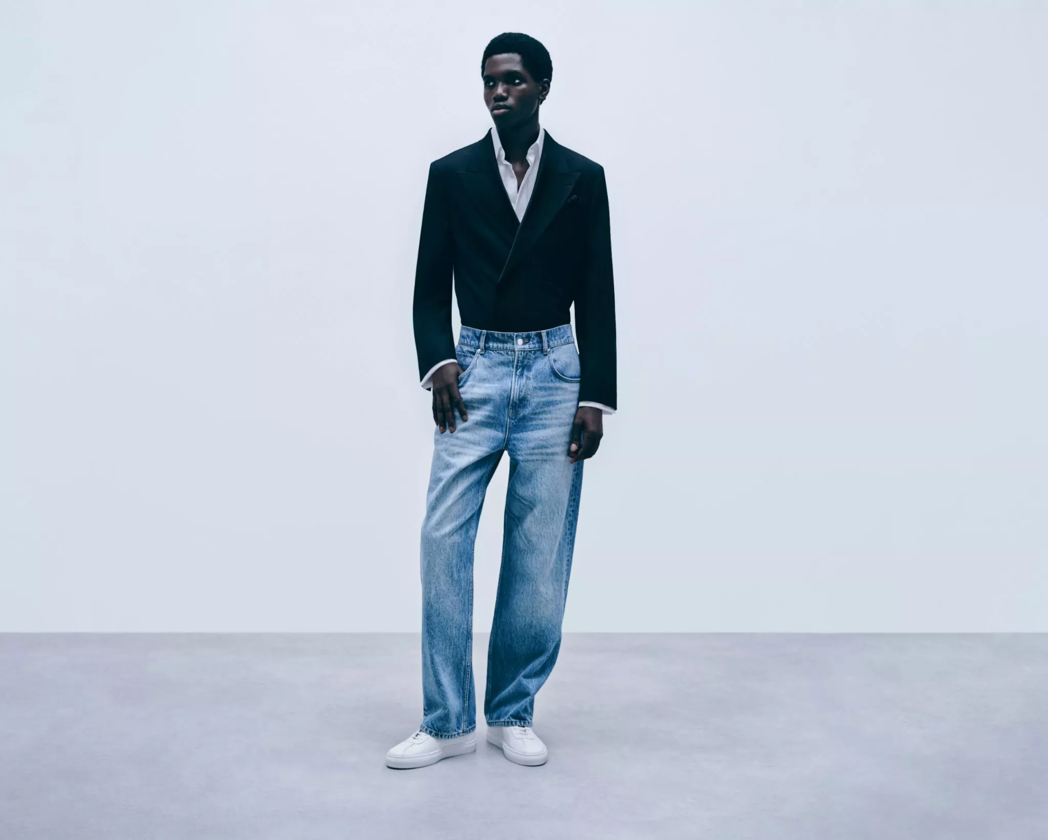 Full-length studio photo of a man in a black blazer, white shirt and wide-leg jeans with white trainers