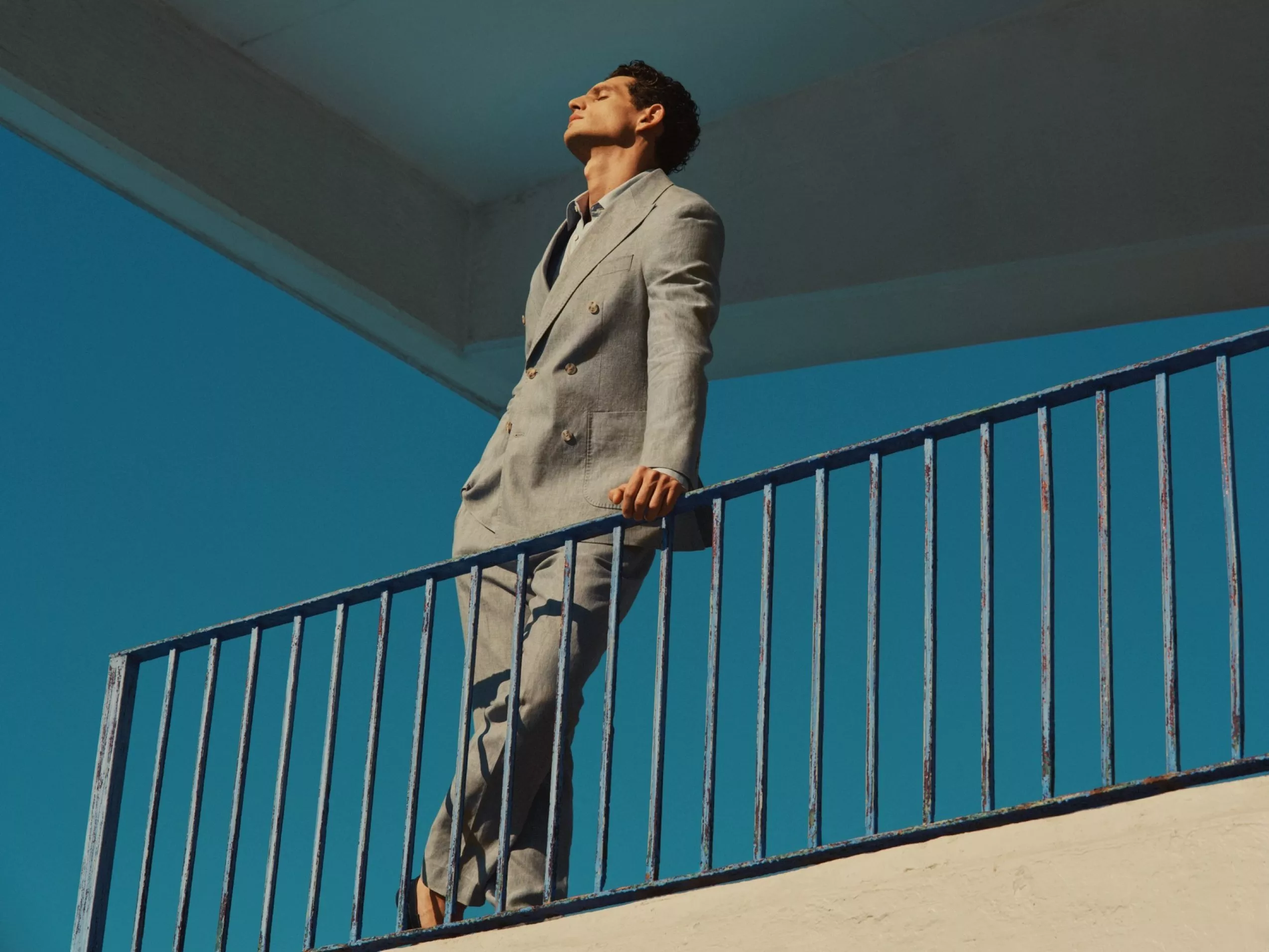 Man in light grey suit standing on outdoor stair railing under blue sky, modern fashion portrait in sunlight