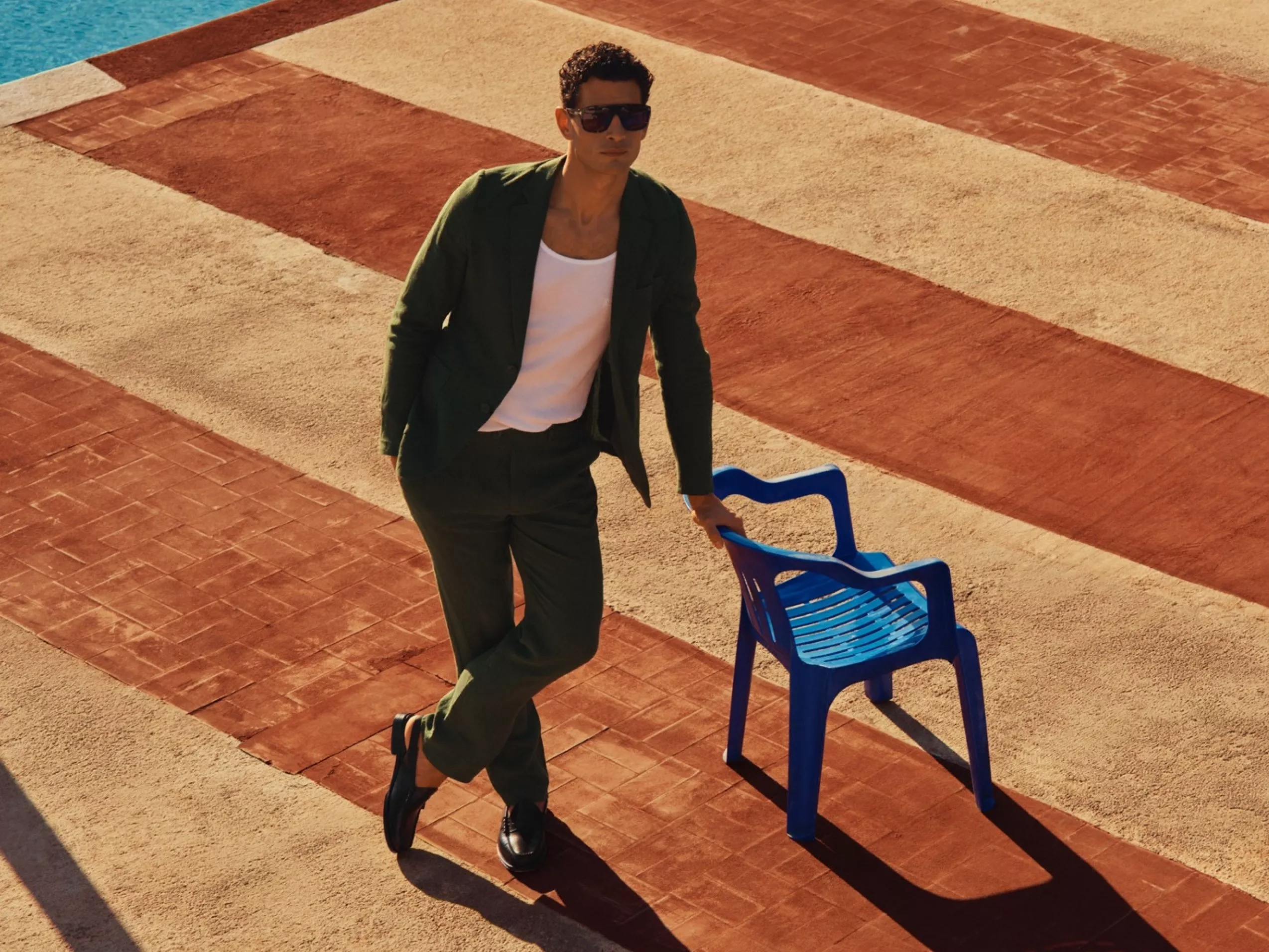 Man in green suit and sunglasses leaning on blue chair by pool, stylish summer menswear fashion photo