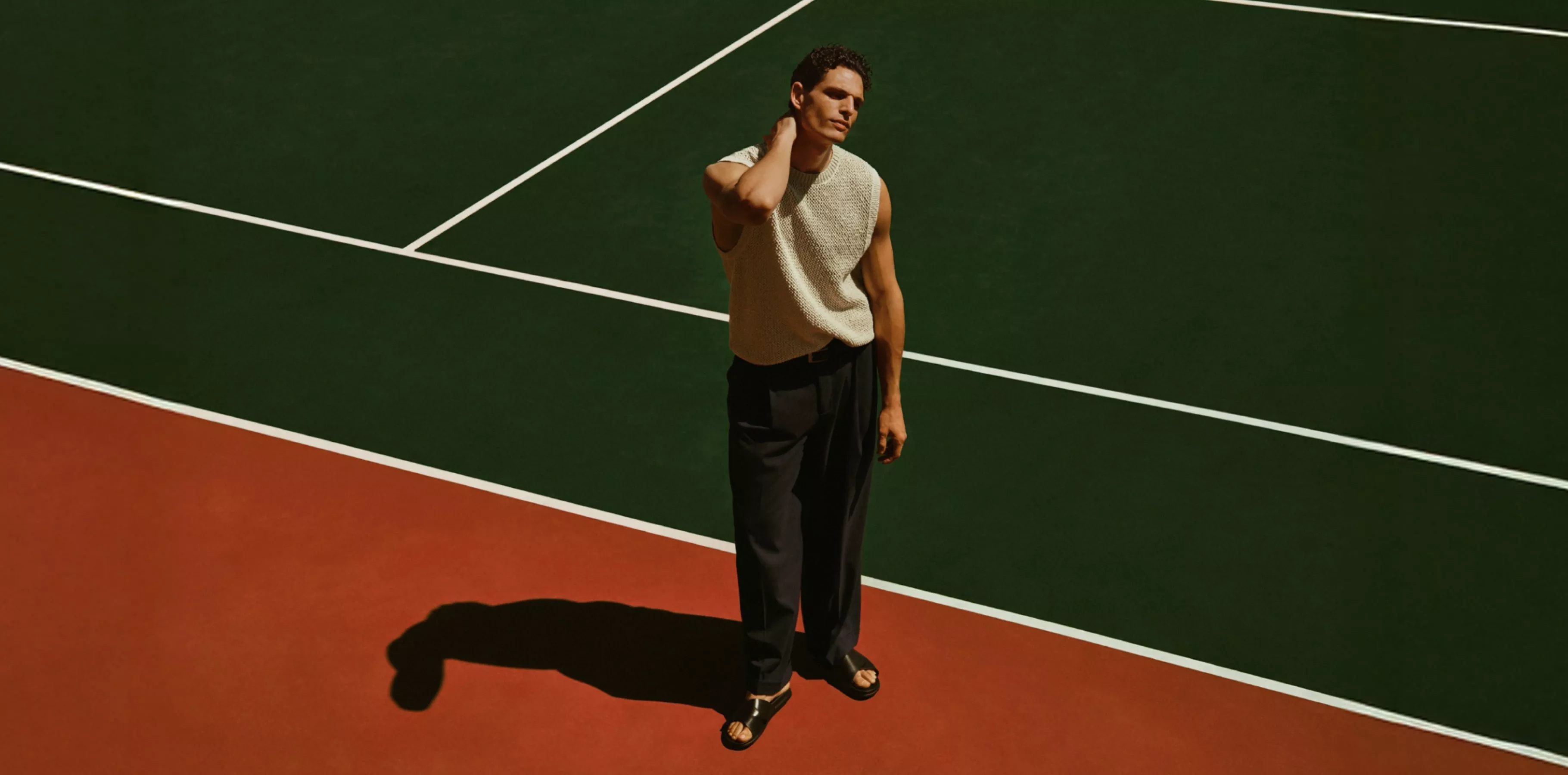 Fashion model in sleeveless knit top and tailored trousers, standing on green and red tennis court lines outdoors