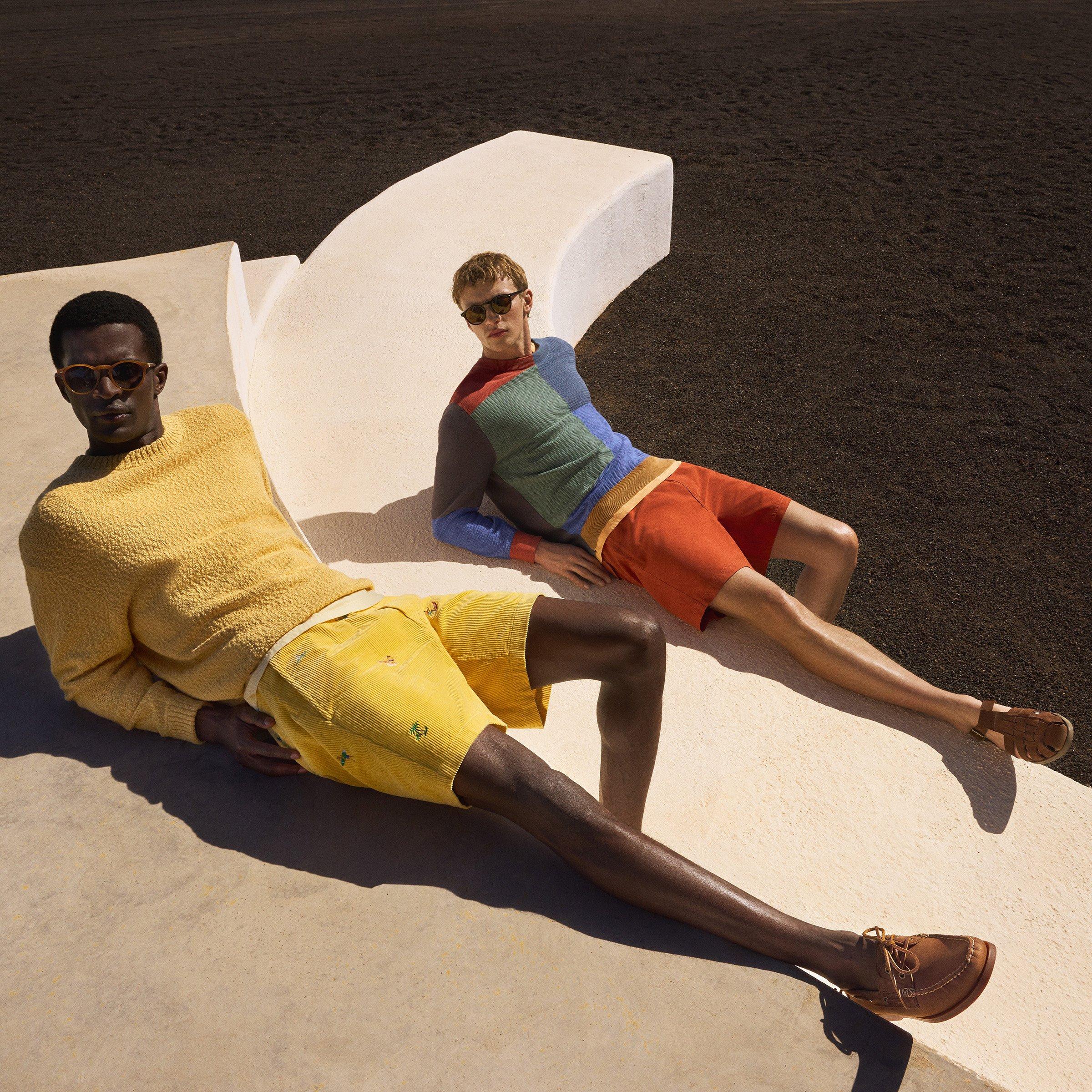 Two men lying across concrete steps, one man in yellow sweater and shorts, the other in mixed block coloured jumper and red shorts.