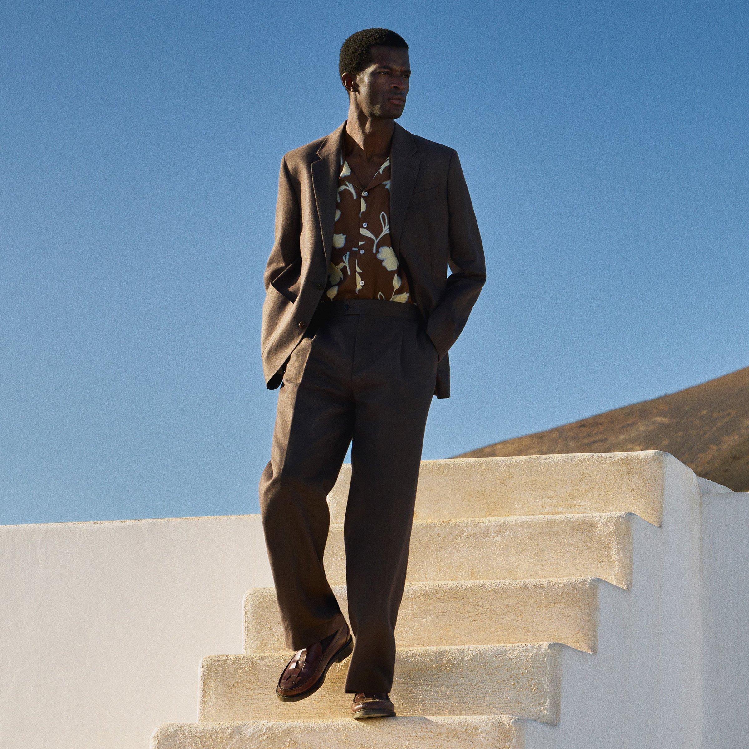 Man in a patterned floral shirt under a beige brown suit jacket and trousers, walking down some steps