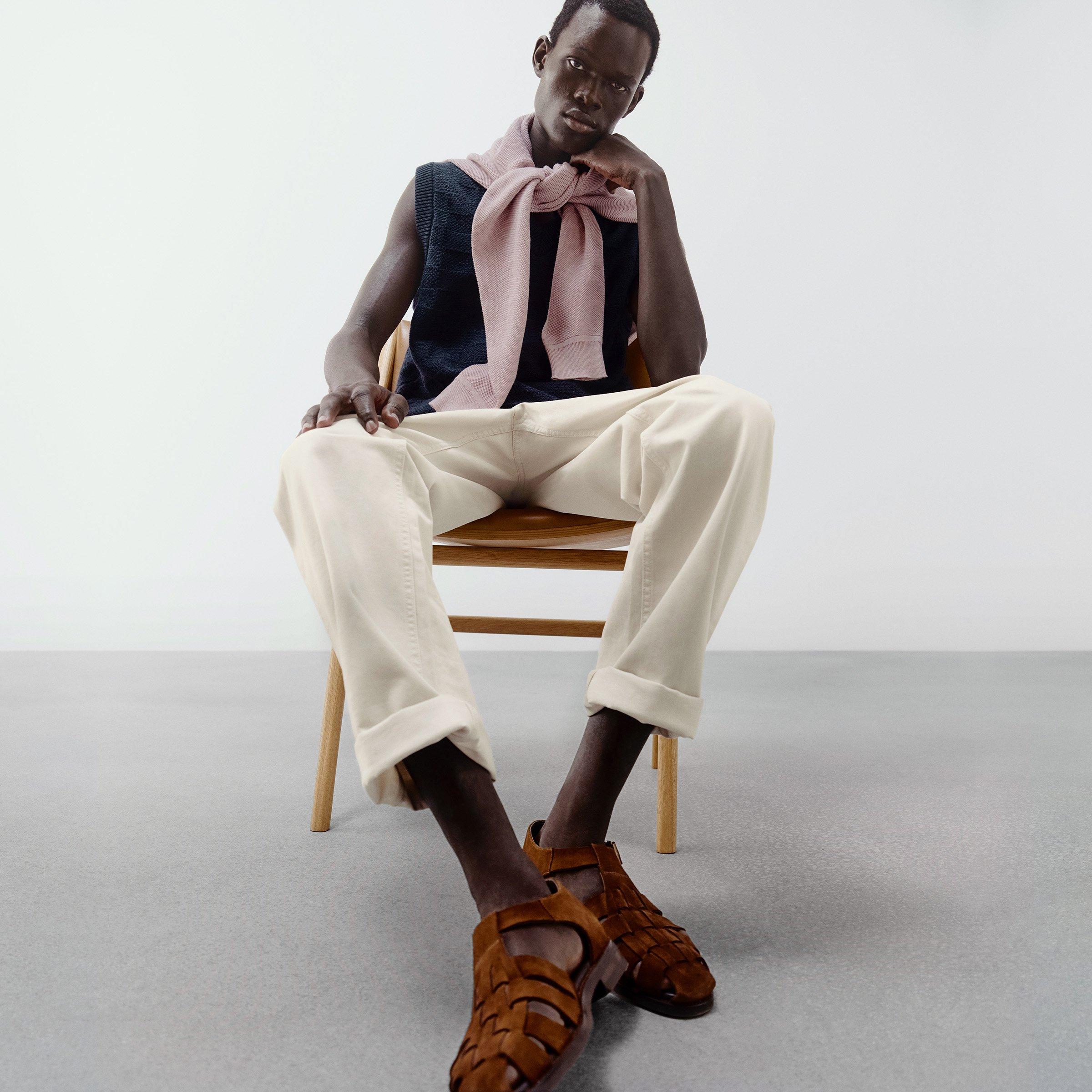 Man sitting on chair in black sleeveless top with pale pink sweater tied around shoulders, cream trousers, and sandals