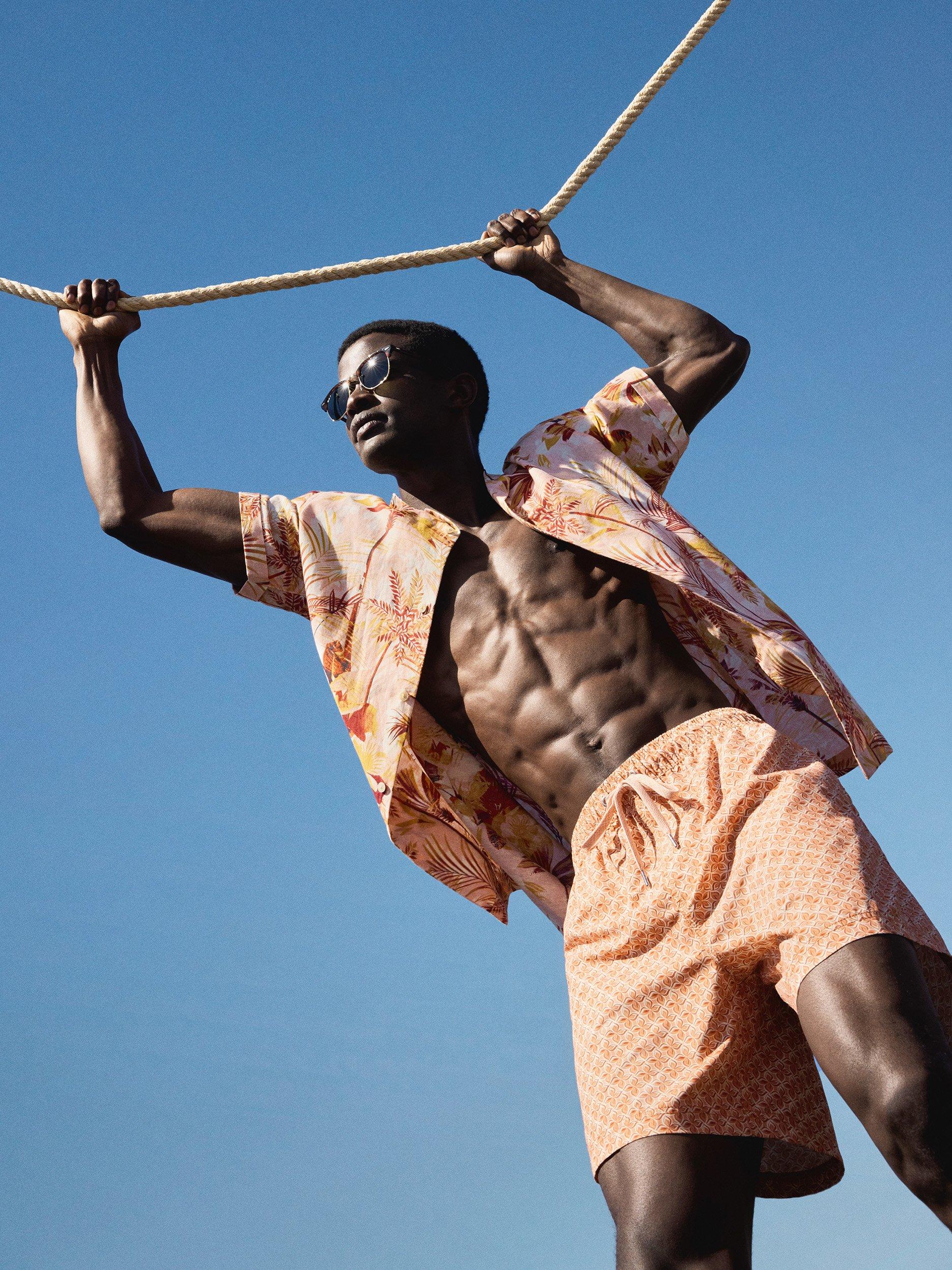 Man holding onto rope, in unbuttoned palm tree print short sleeved shirt, and shorts