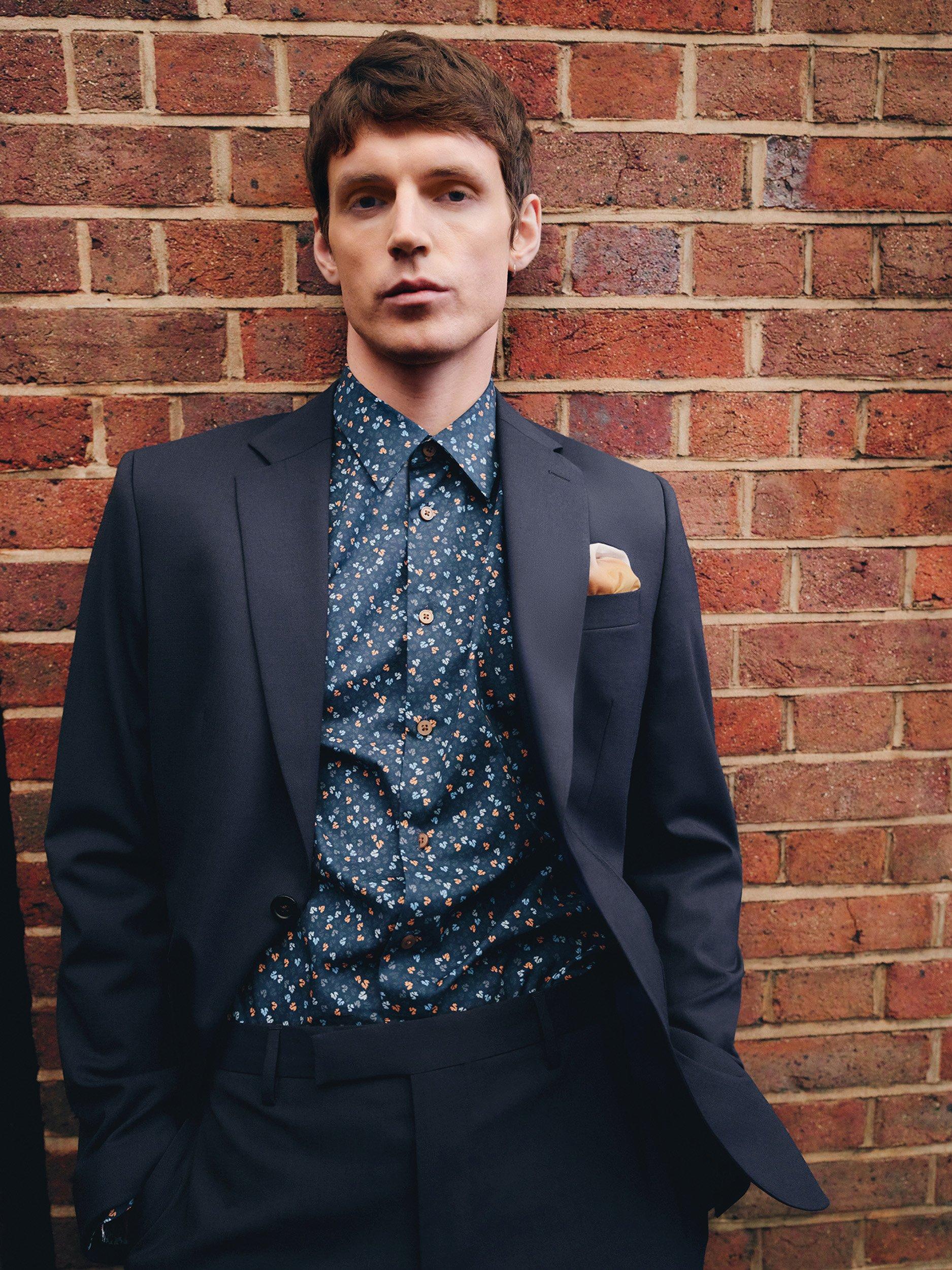 Man leaning against brick wall, wearing spotty blue shirt underneath navy blue suit jacket and trousers, lea