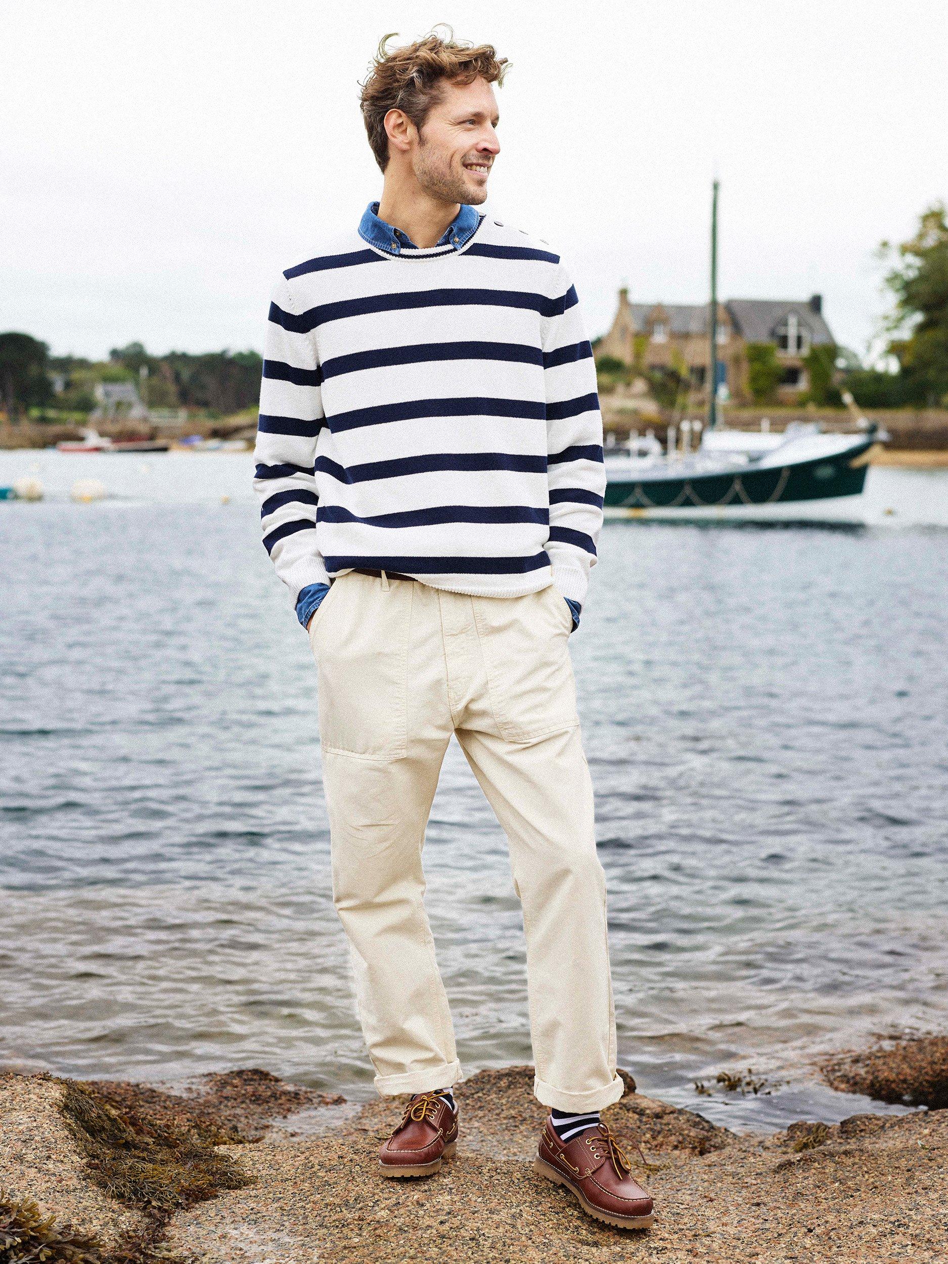 Man standing on sandy bay next to water, wearing navy blue and white striped jumper, khaki trousers and leather loafers