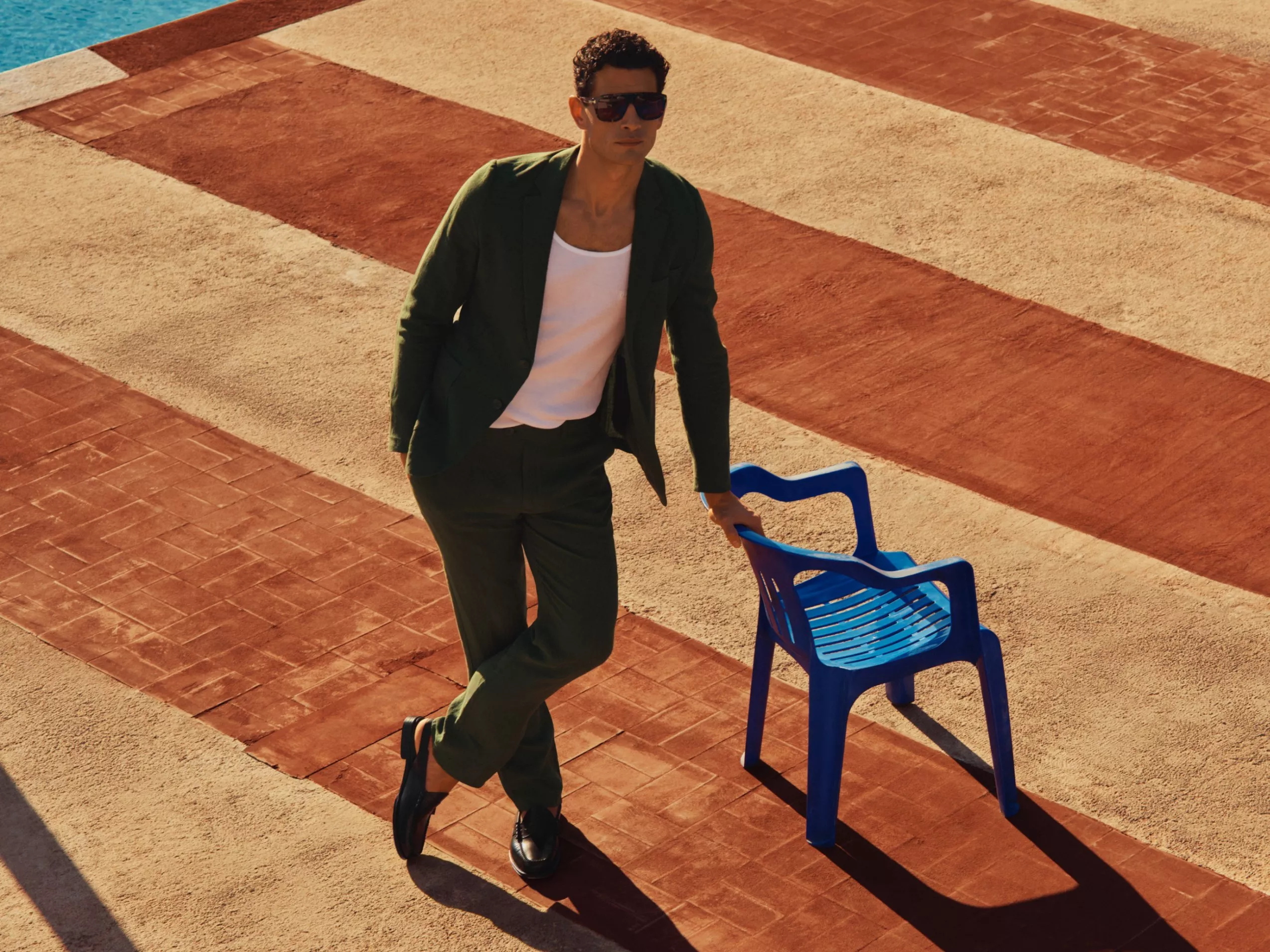 Man in green suit leaning on blue plastic chair by poolside, stylish summer fashion portrait in sunlight