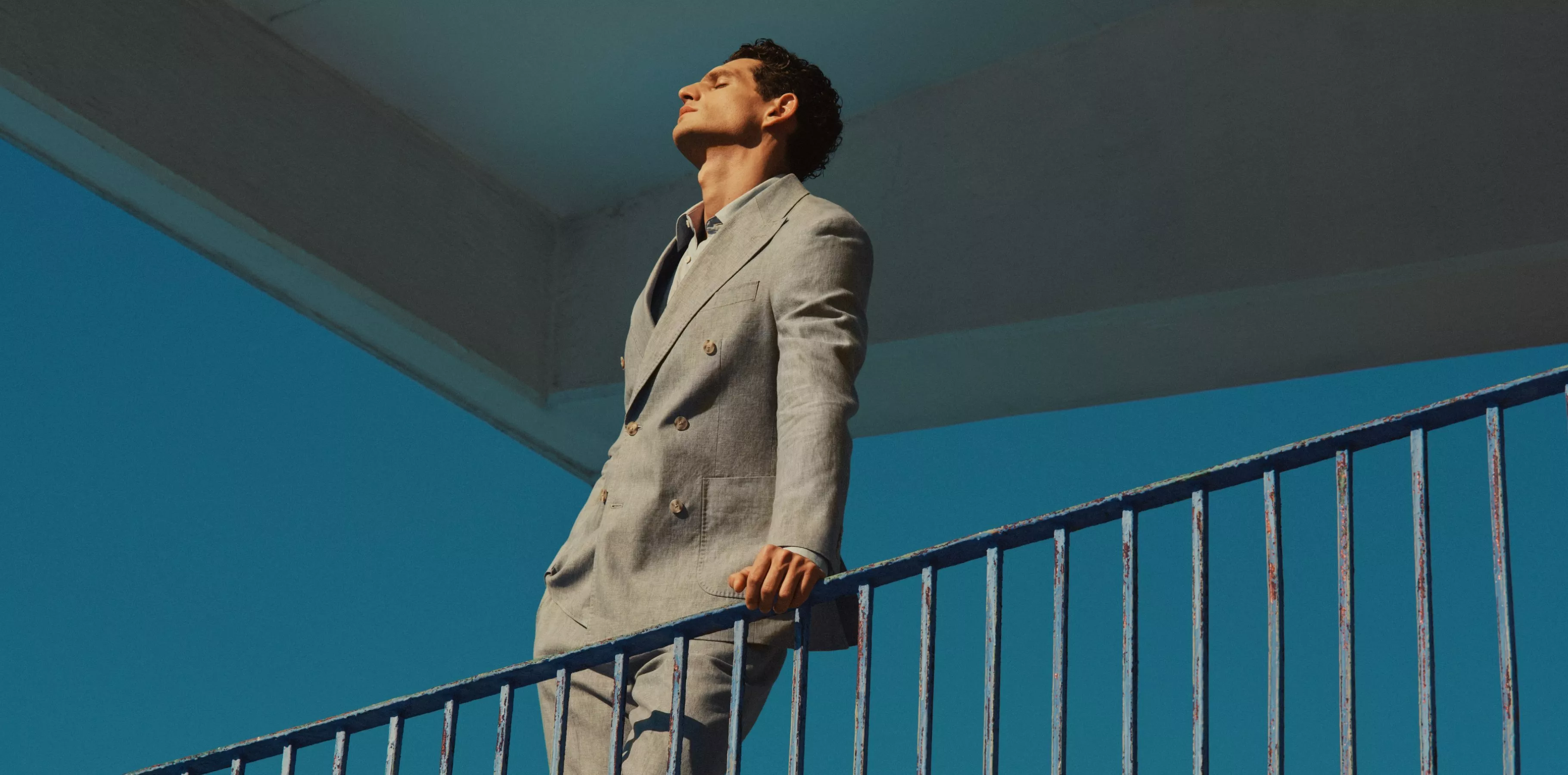 man wearing a grey linen suit with his hand holding a blue railing