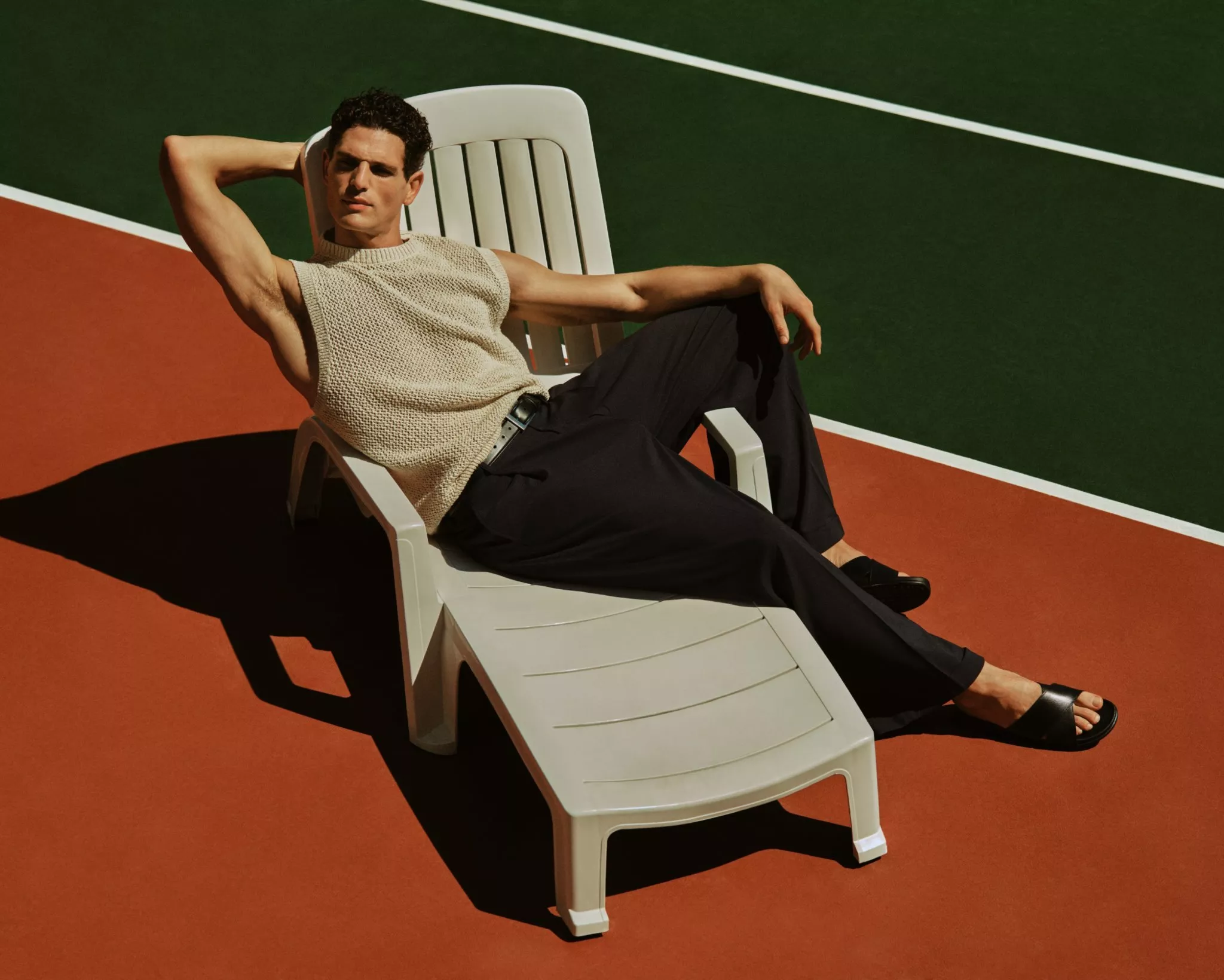 Male model on a sun lounger beside a tennis court, wearing a knitted vest, black trousers and black sliders