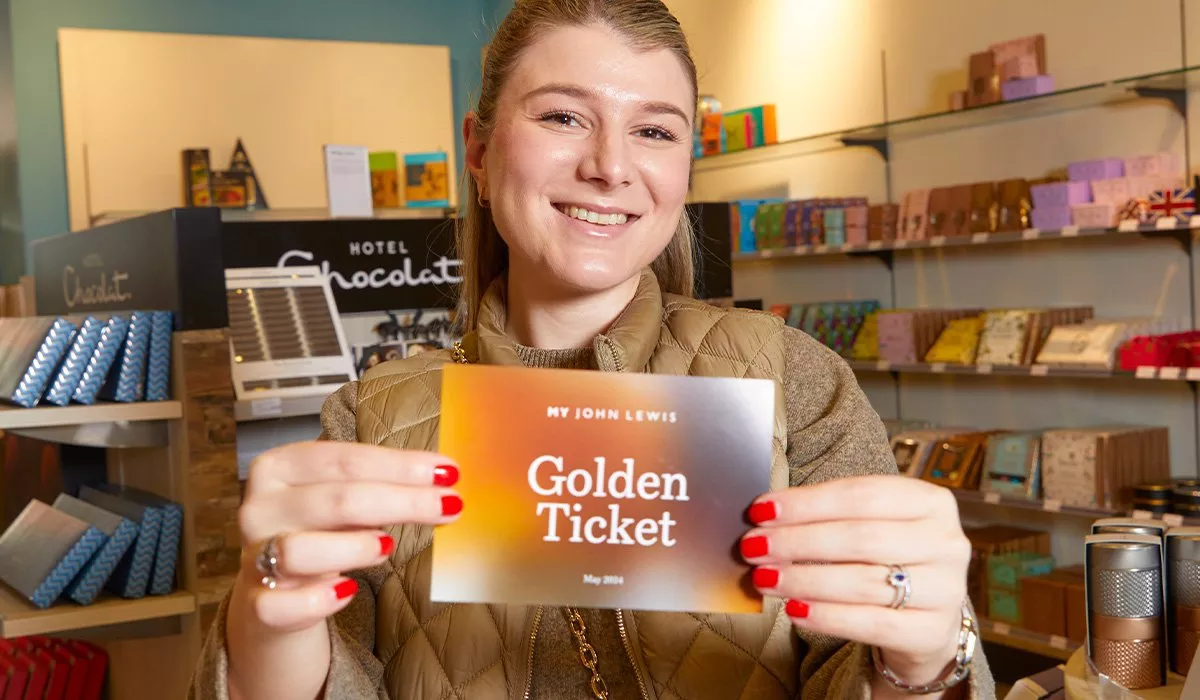Smiling woman holding a John Lewis Golden Ticket in a chocolate shop, with shelves of luxury chocolates behind.