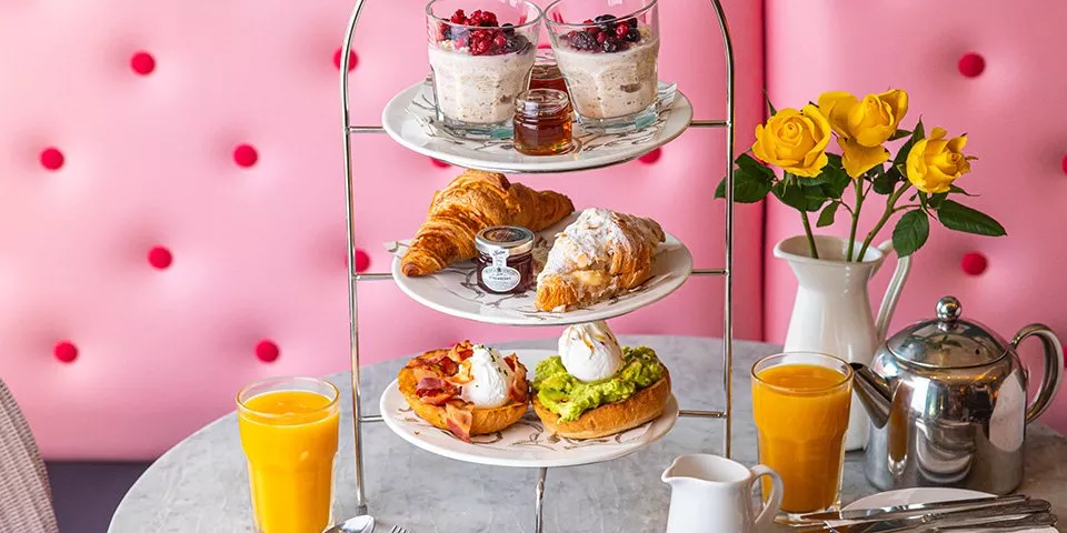 A tiered stand with croissants, avocado toast, overnight oats, and orange juice on a table with yellow roses.