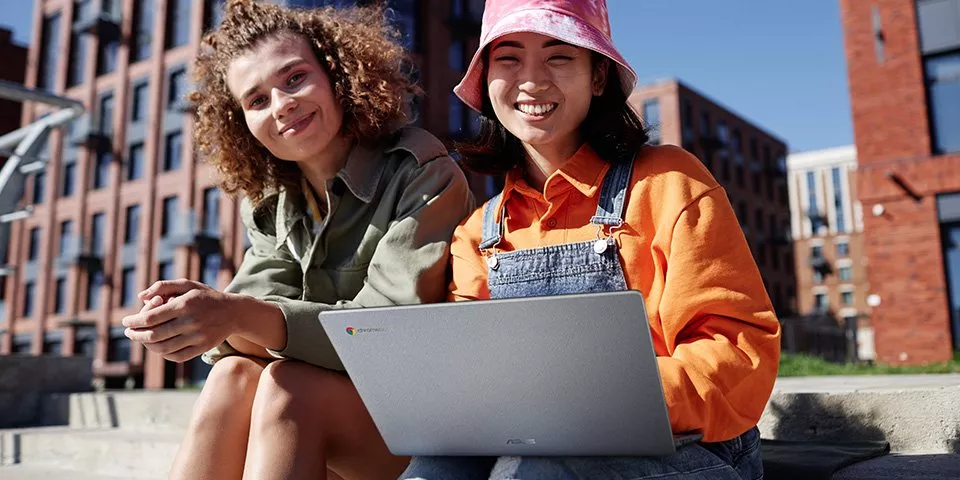 Two smiling friends using a Chromebook laptop outdoors in a modern city courtyard on a sunny day