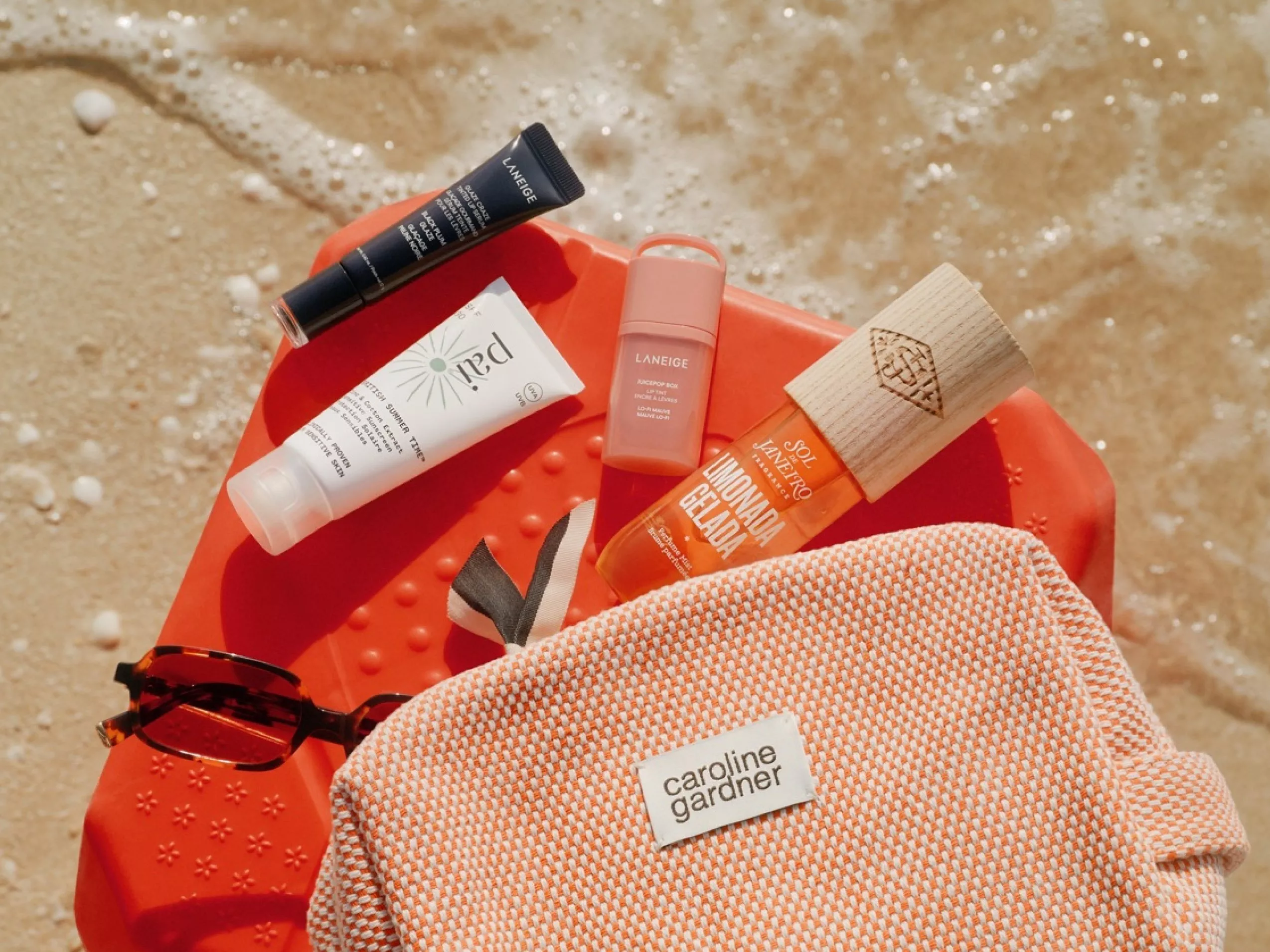 Beach flat lay with sunscreen, lip tint, sunglasses and tote bag on orange tray by the sea foam shoreline