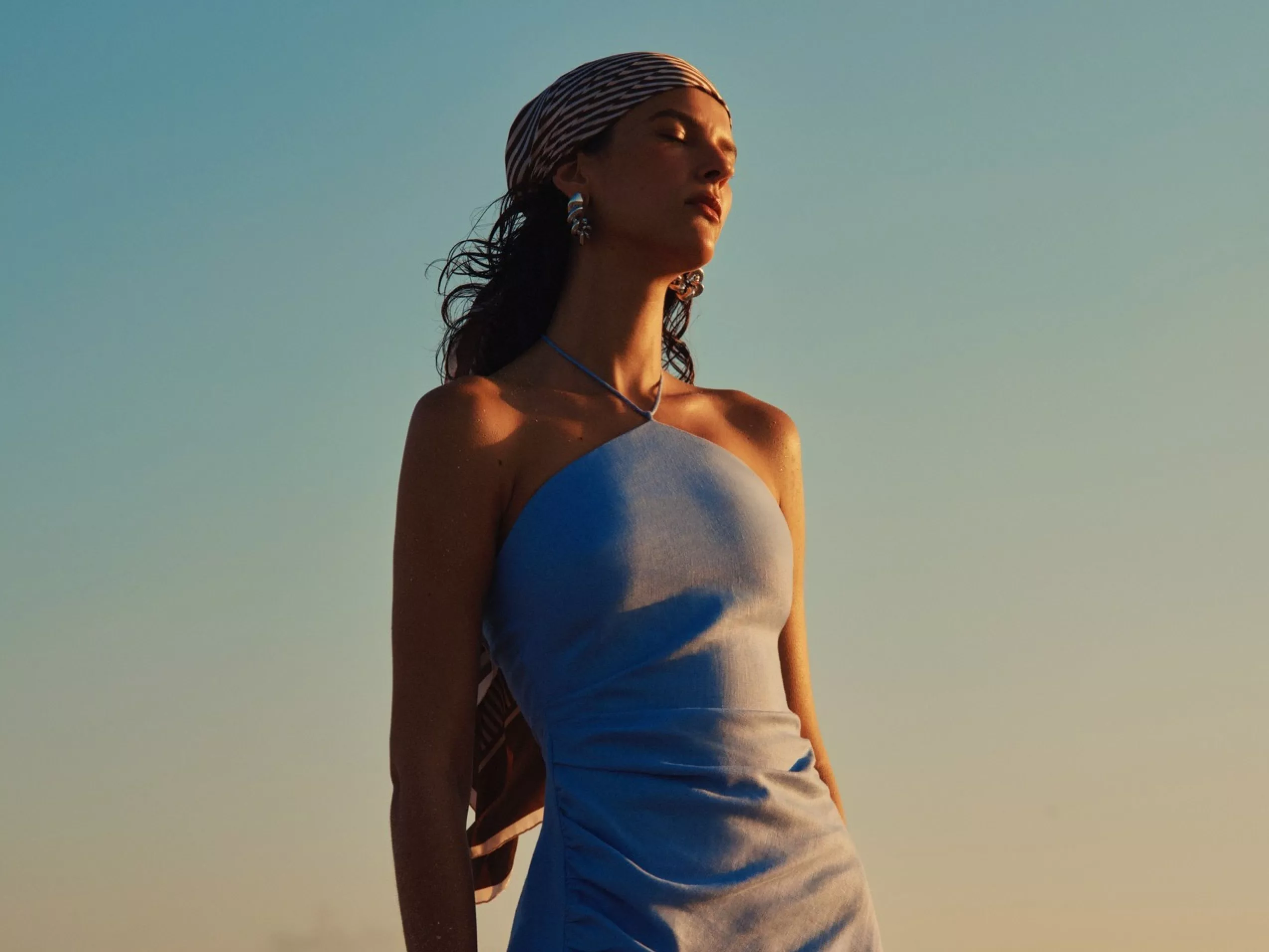 Woman in light blue halter dress with headscarf, posing outdoors in golden hour sunset light against clear sky