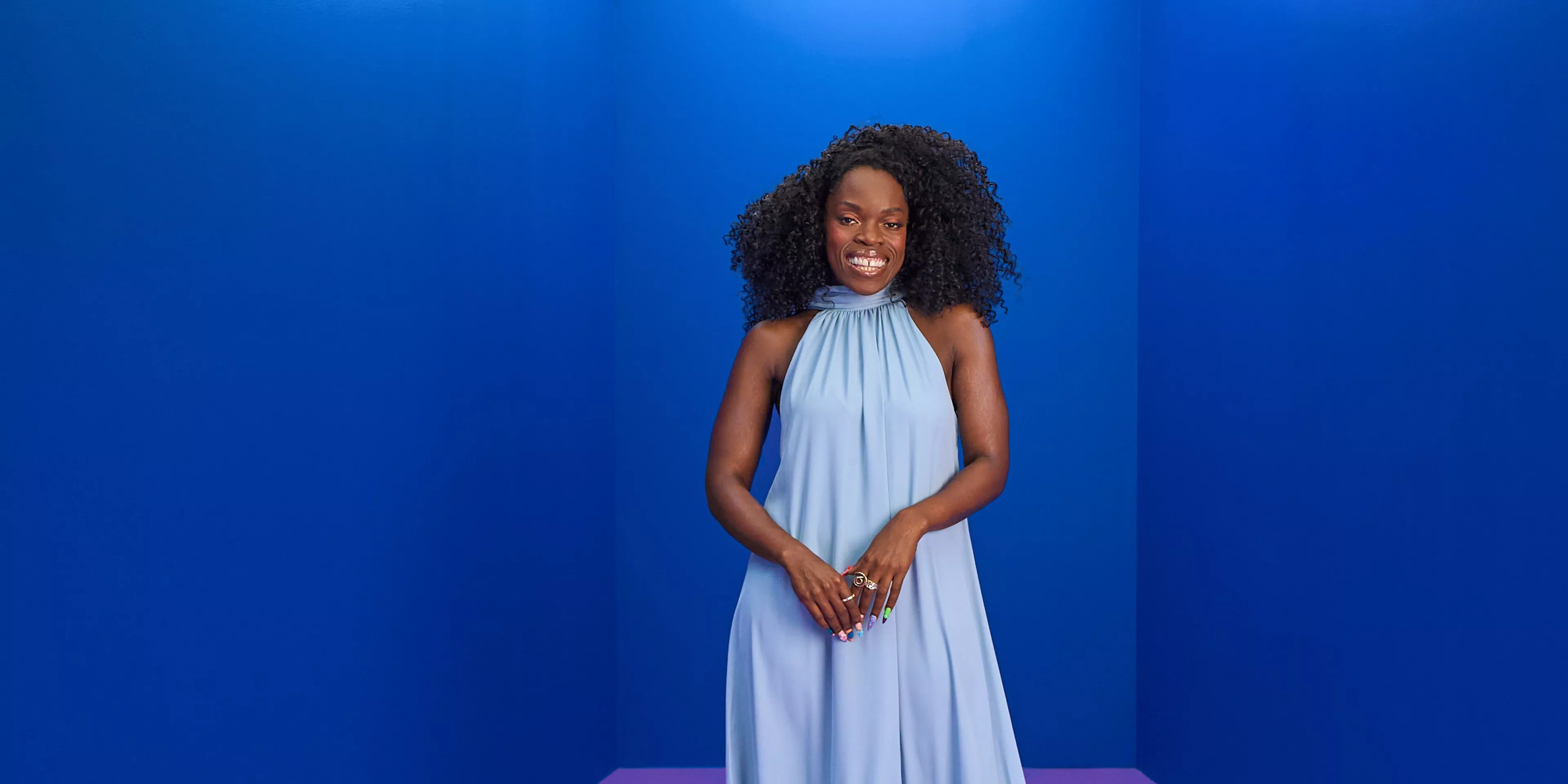 Smiling Nana Acheampong with curly hair in a light blue halter dress, standing against a bold blue studio backdrop