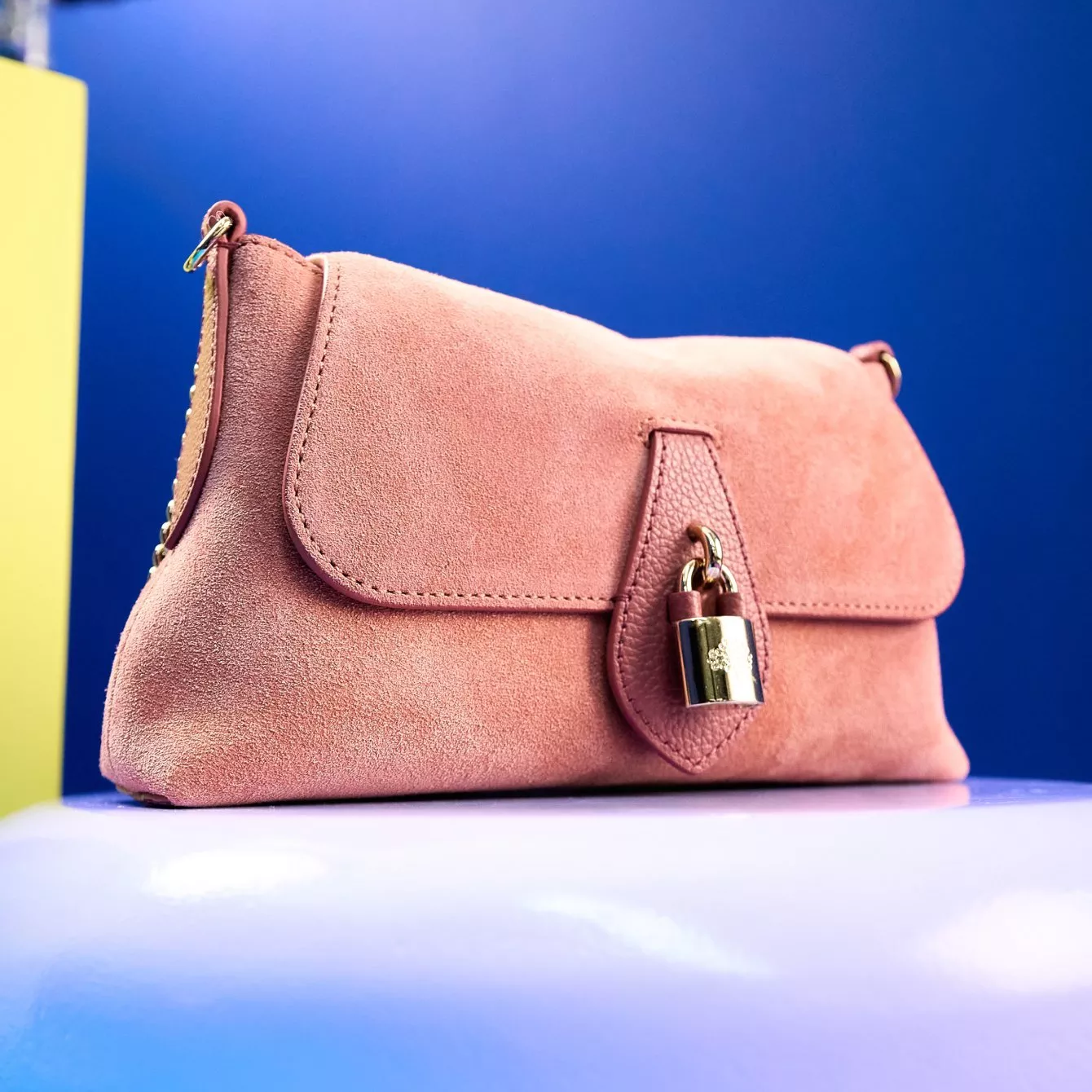 Blush pink suede crossbody Mulberry handbag with gold padlock detail, photographed on a blue background for fashion retail.