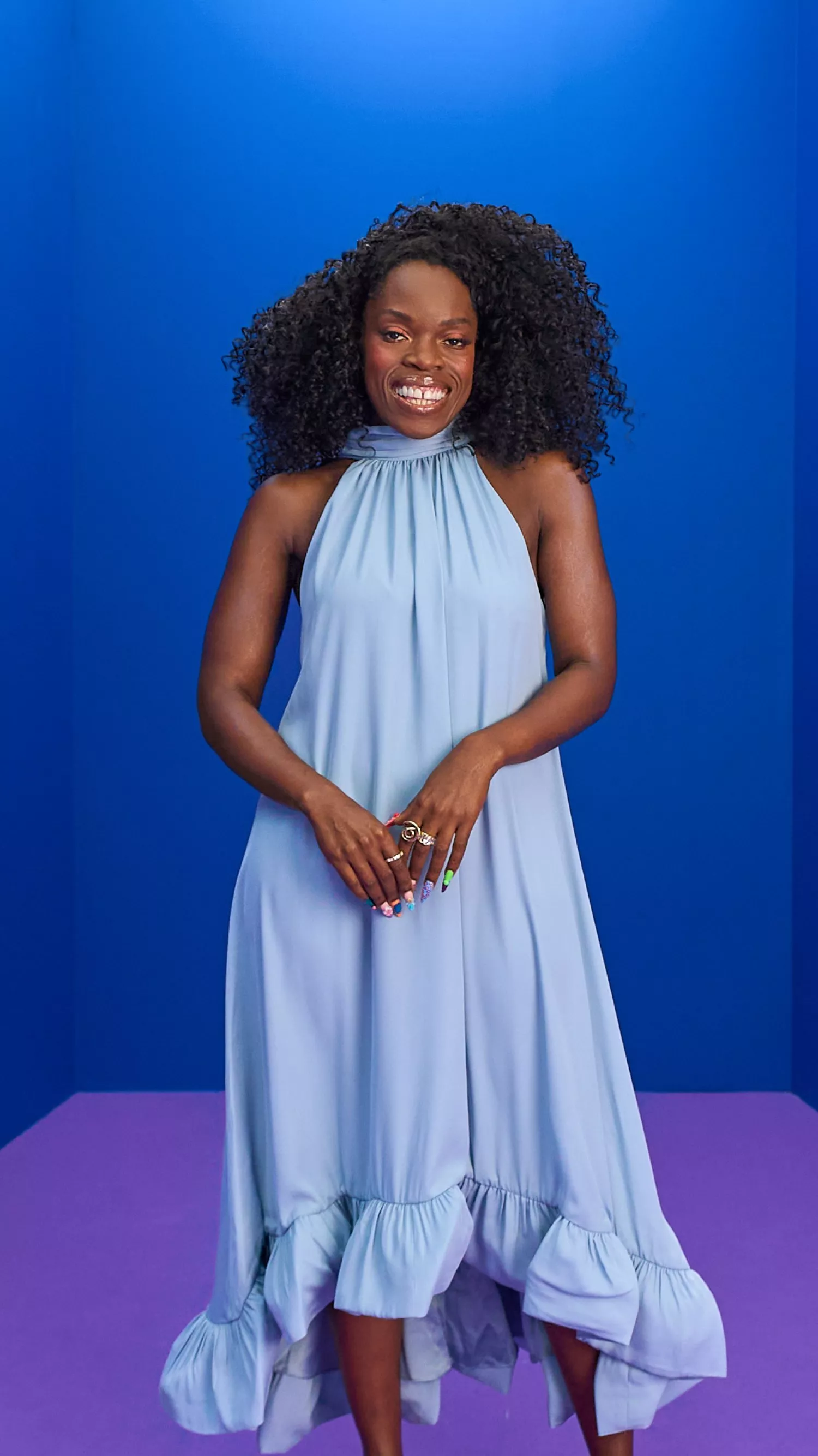 Smiling Nana Acheampong with curly hair in a light blue halter dress, standing against a bold blue studio backdrop