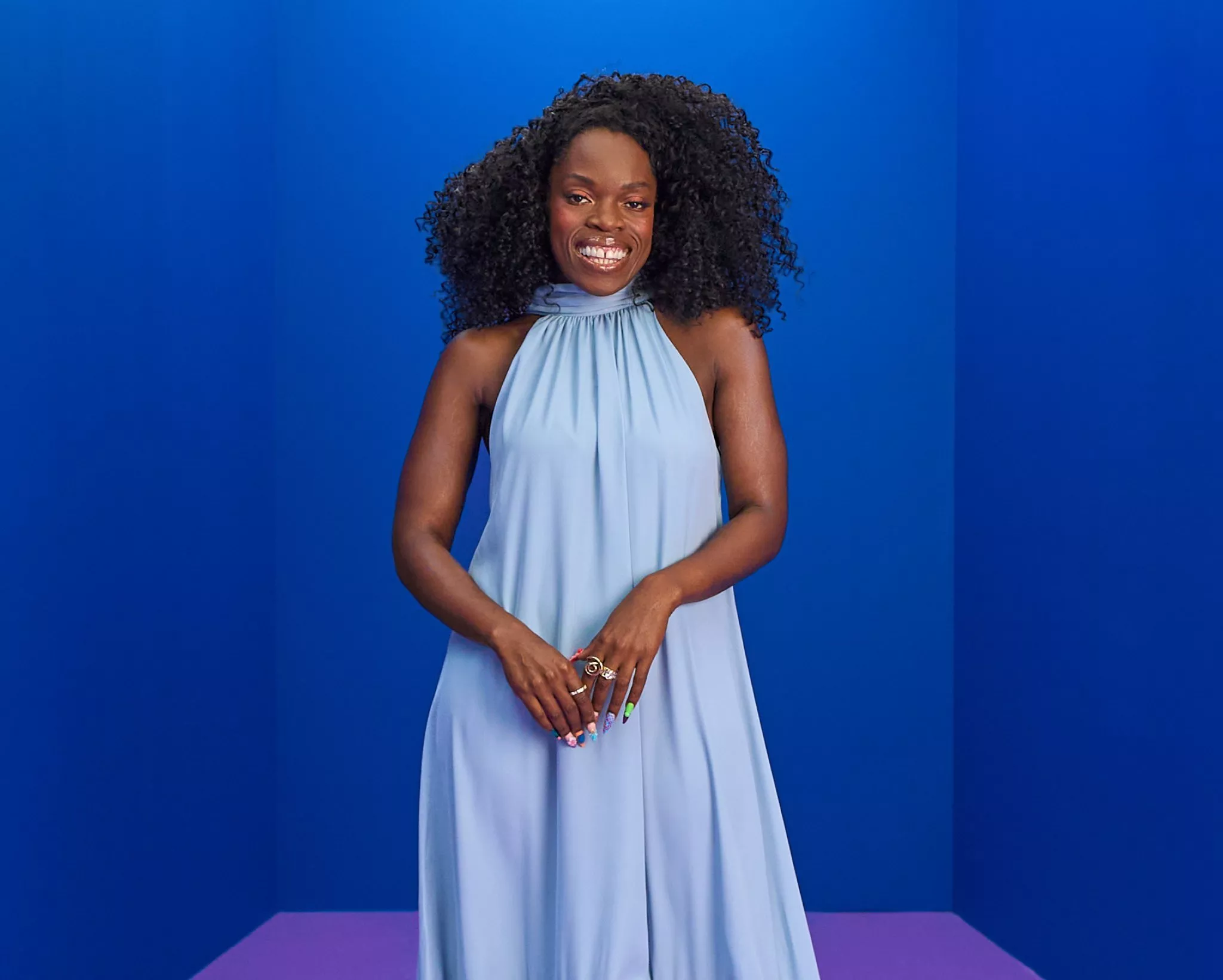 Smiling Nana Acheampong with curly hair in a light blue halterneck maxi dress, posing in a bright blue studio backdrop