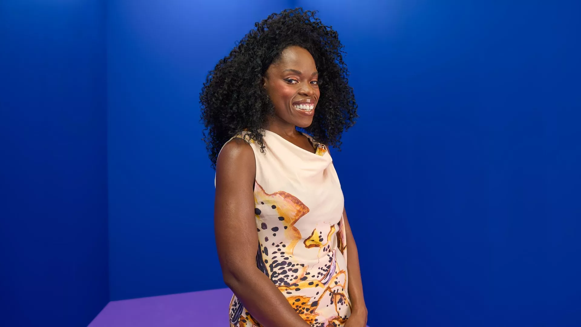 Portrait of Nana Acheampong with curly hair in sleeveless patterned dress, standing in blue studio background
