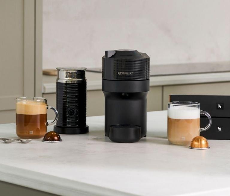 Black Nespresso coffee machine and milk frother on a kitchen counter with coffee pods, capsule boxes, and two glass mugs of coffee.