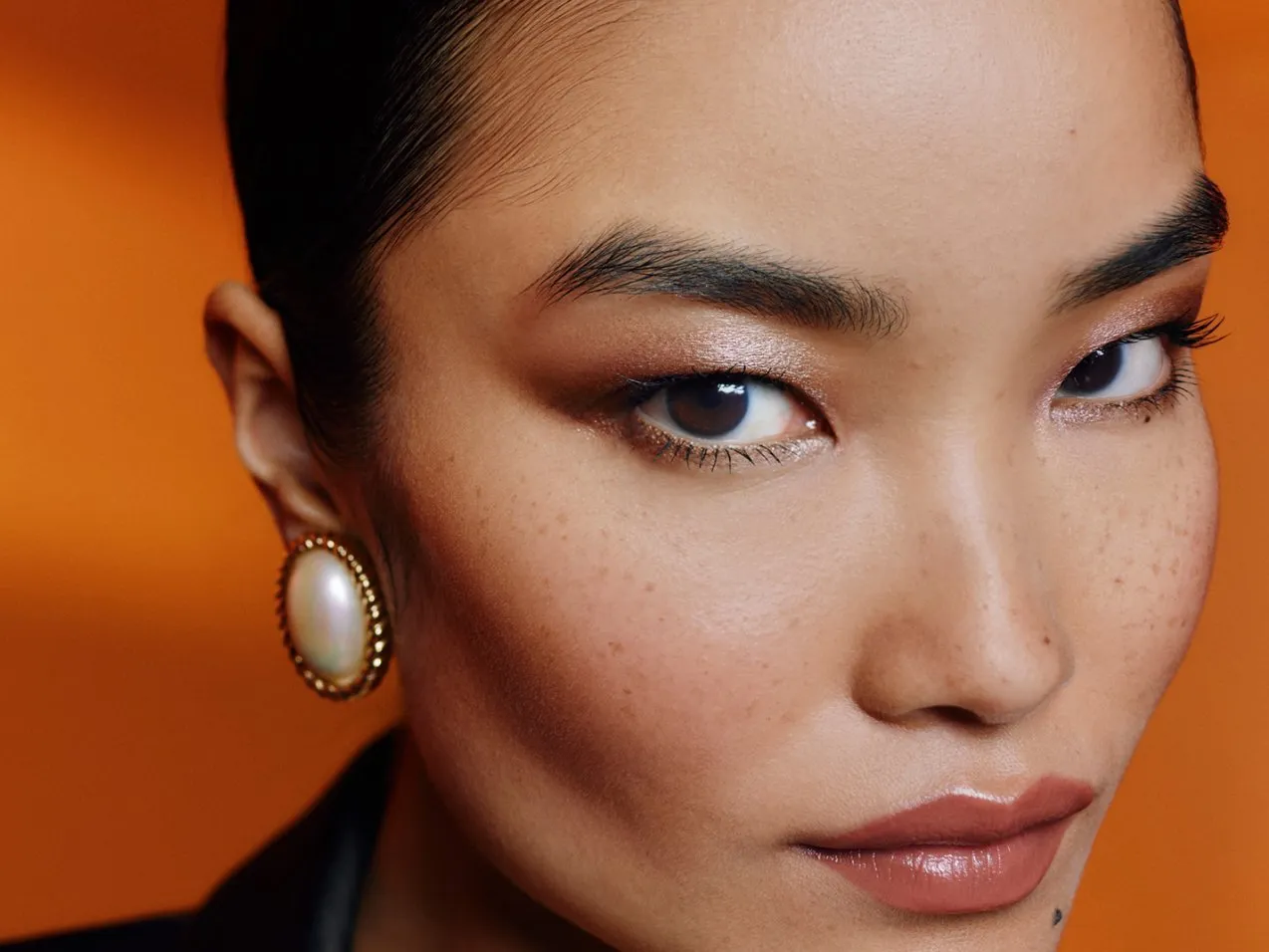 Close-up portrait of woman with natural freckles, glossy lips, pearl earring and warm bronze eye makeup on orange background
