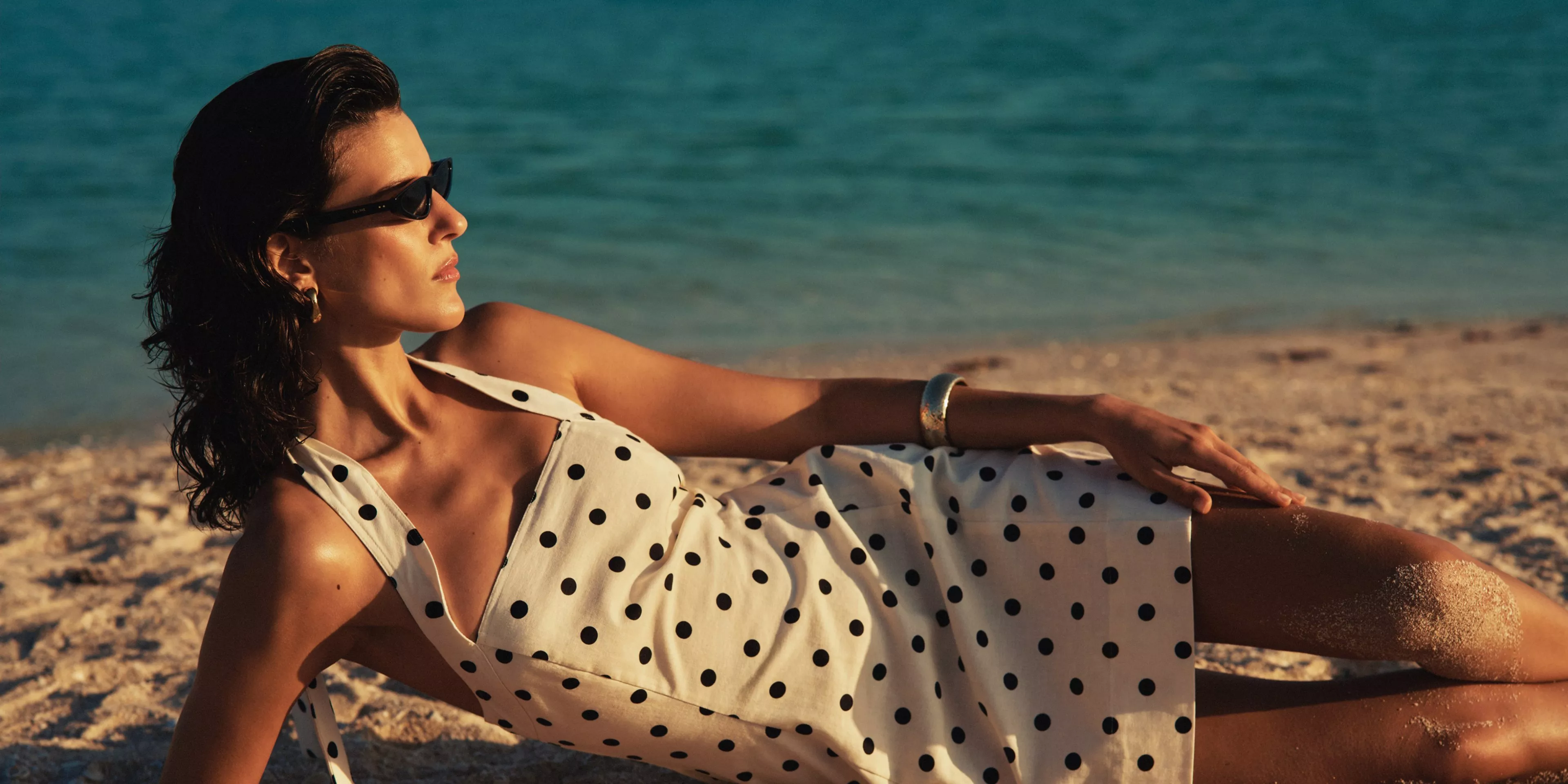 Woman in a polka dot dress and sunglasses reclining on sandy beach by the sea in warm sunlight