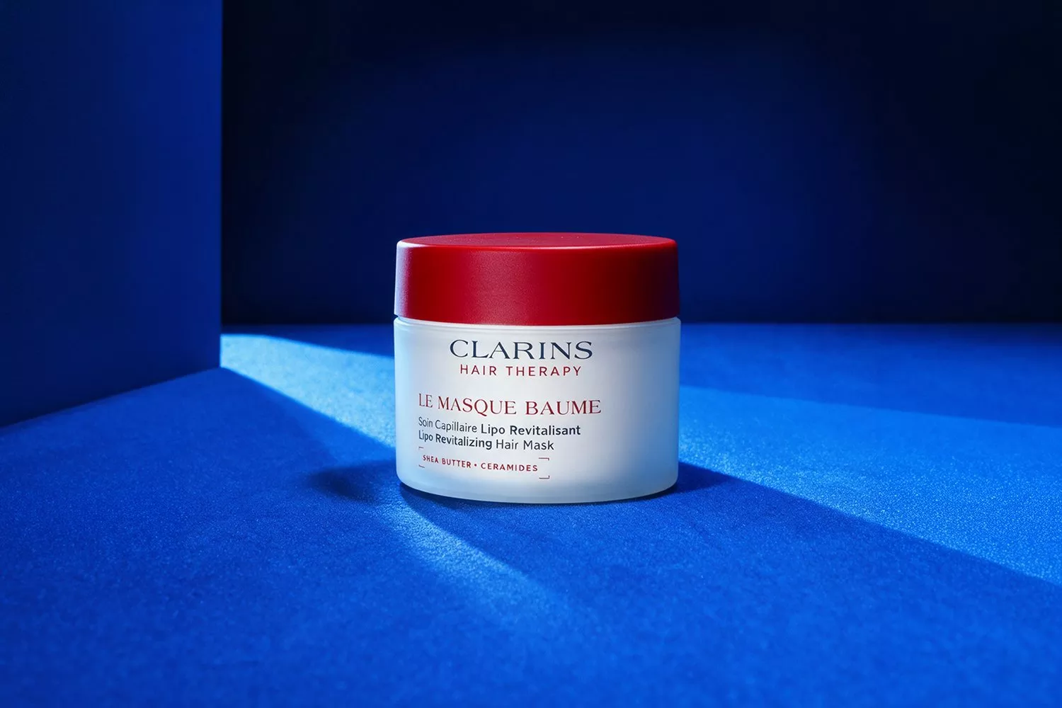 Clarins Hair Therapy Le Masque Baume revitalising hair mask jar with red lid on blue background in studio light