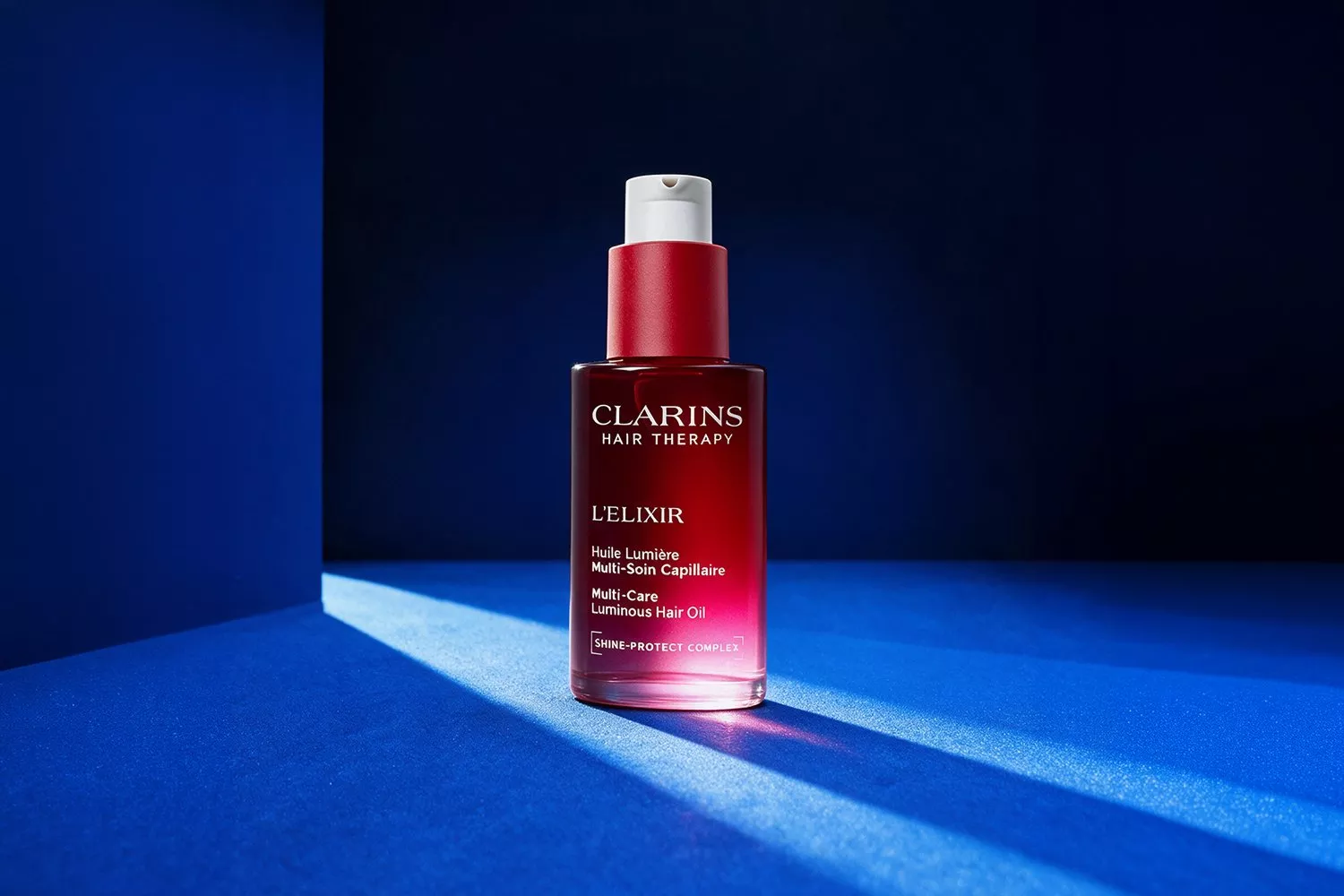 Clarins Hair Therapy L’Elixir luminous hair oil bottle on blue studio backdrop with dramatic light and shadow.