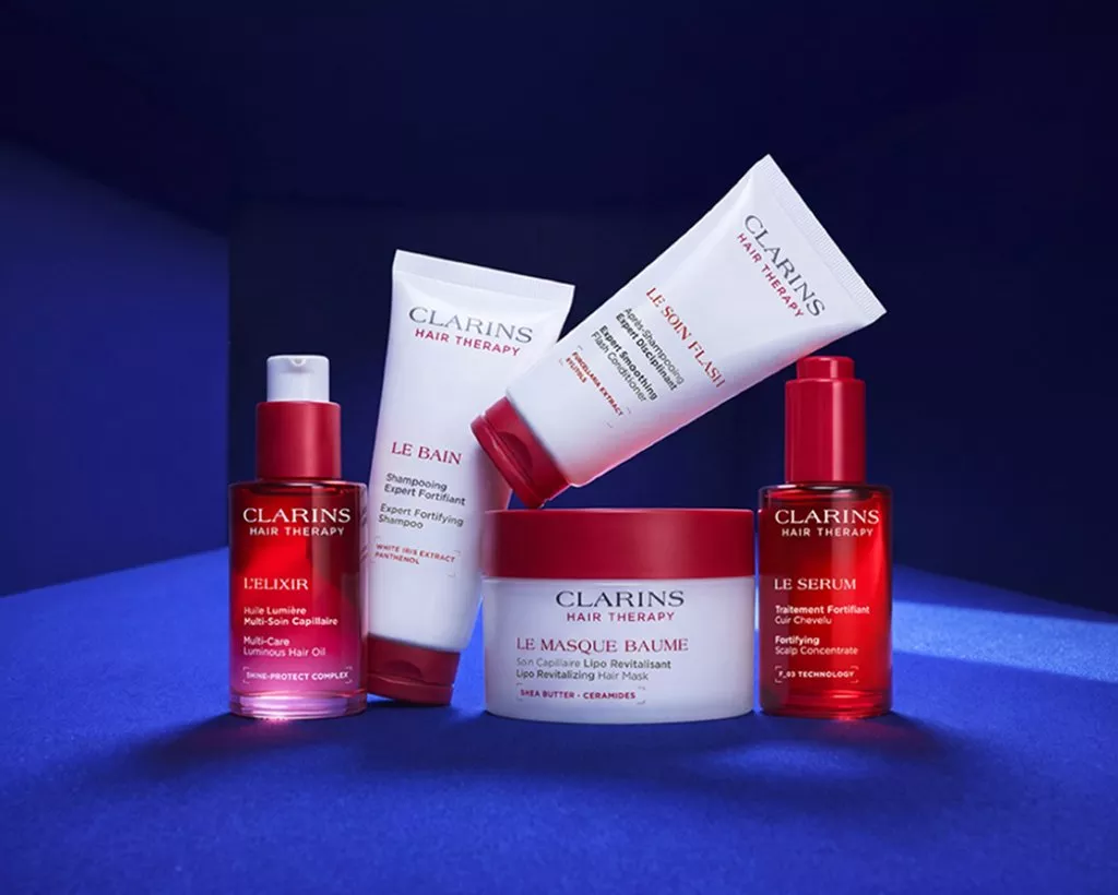 Clarins Hair Therapy products on blue background: shampoo, conditioner, hair mask jar, serum and hair oil bottles.