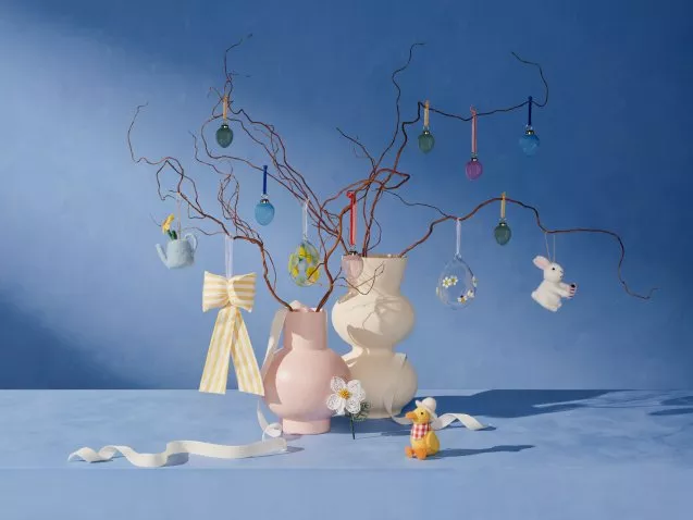 Decorative branches in pink and white vases with hanging Easter ornaments, a ribbon, and a toy duck on a blue backdrop.