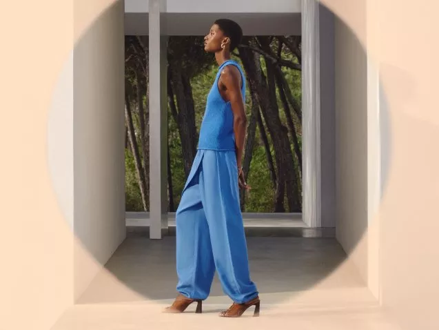 A person in stylish blue attire walks confidently, framed by a modern architectural setting with trees visible outside.