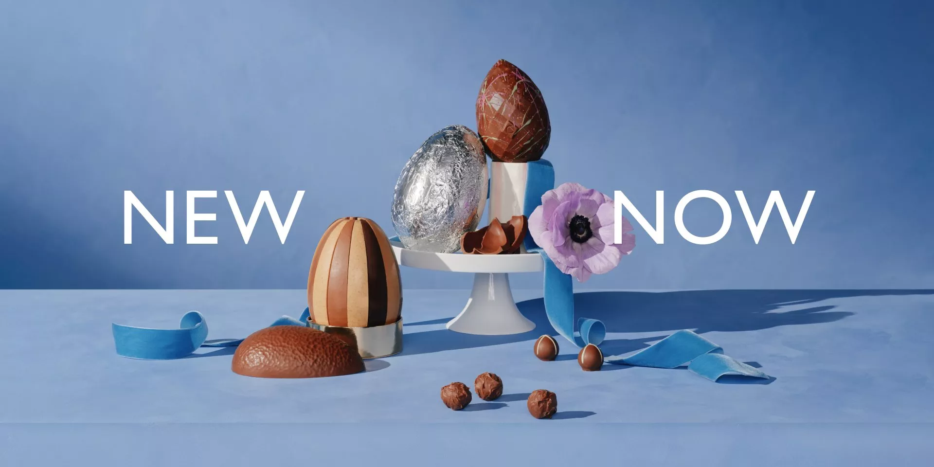 Elegant display of chocolate eggs with vibrant blue ribbon and flower on a cake stand against a blue backdrop.