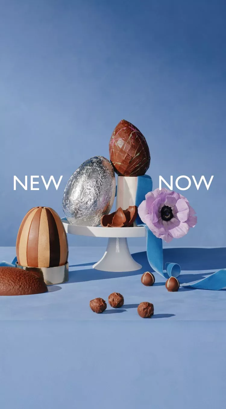 Assorted decorative chocolate eggs on a stand with a purple flower and ribbon, set against a blue backdrop.
