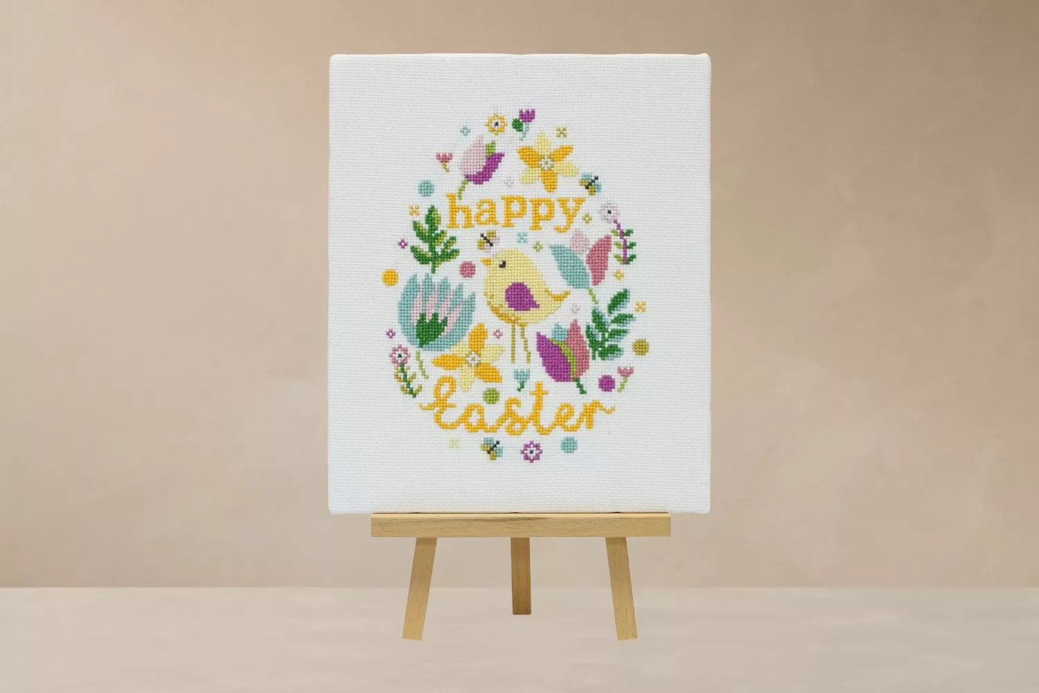 A decorative Easter embroidery on a small easel, featuring flowers, a bird, and the words "Happy Easter".