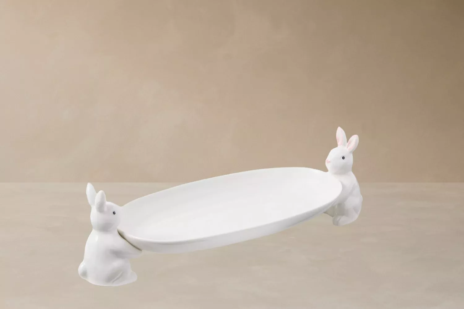 White oval plate with two ceramic bunny figurines holding it on a light beige background.